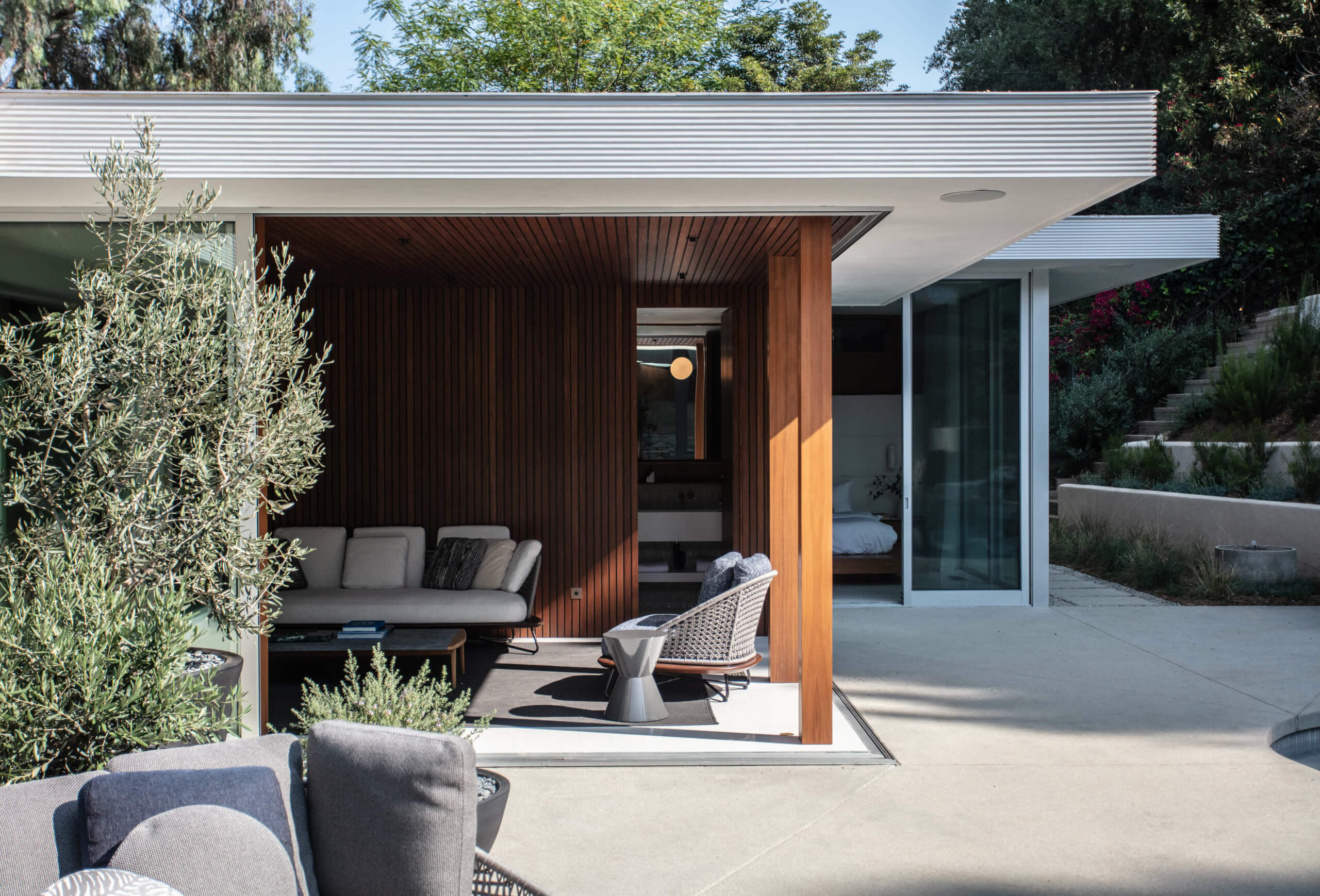 Mid-Century Modern Restoration — Modern residential pavilion with dark timber cladding, concrete terrace, and integrated seating area surrounded by olive trees and minimalist landscaping.
