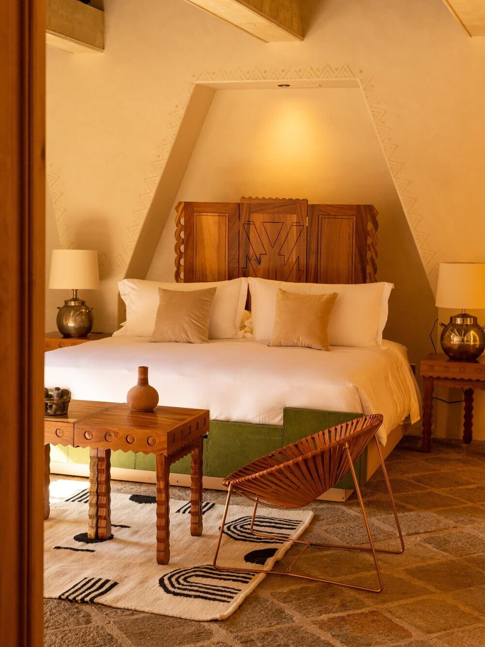 La Valise San Miguel, Mexico — Ochre bedroom with carved wooden headboard, brass lamps, geometric-patterned rugs, and modernist lounge chair beneath sloped ceiling.