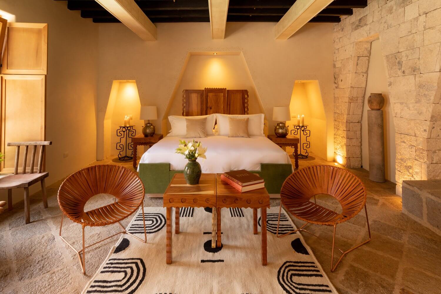La Valise San Miguel, Mexico — Ochre-walled bedroom with exposed wooden beams, terracotta lounge chairs, stone accent wall, warm ambient lighting, and minimalist mid-century furnishings.