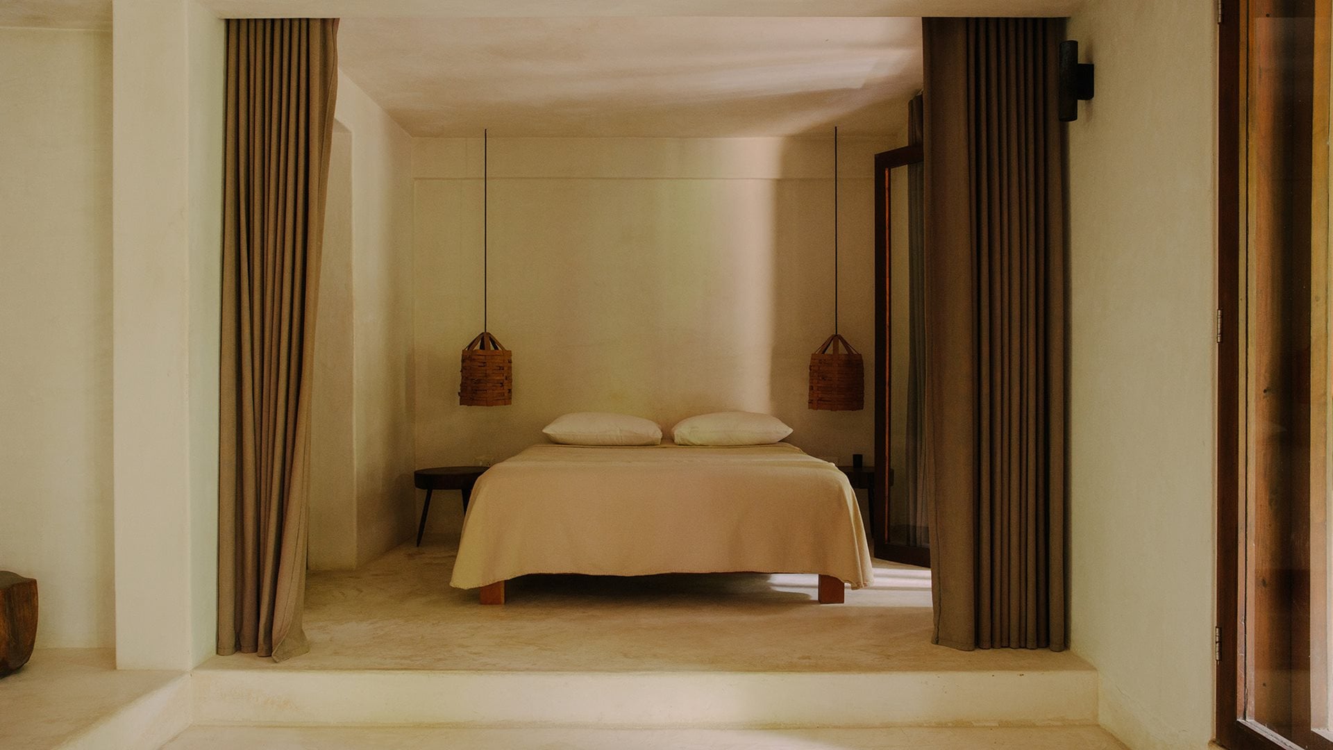 Tulum Treehouse, Mexico — Minimalist bedroom with raised platform bed, cream bedding, woven pendant lights, floor-to-ceiling curtains, and warm natural light filtering through openings.