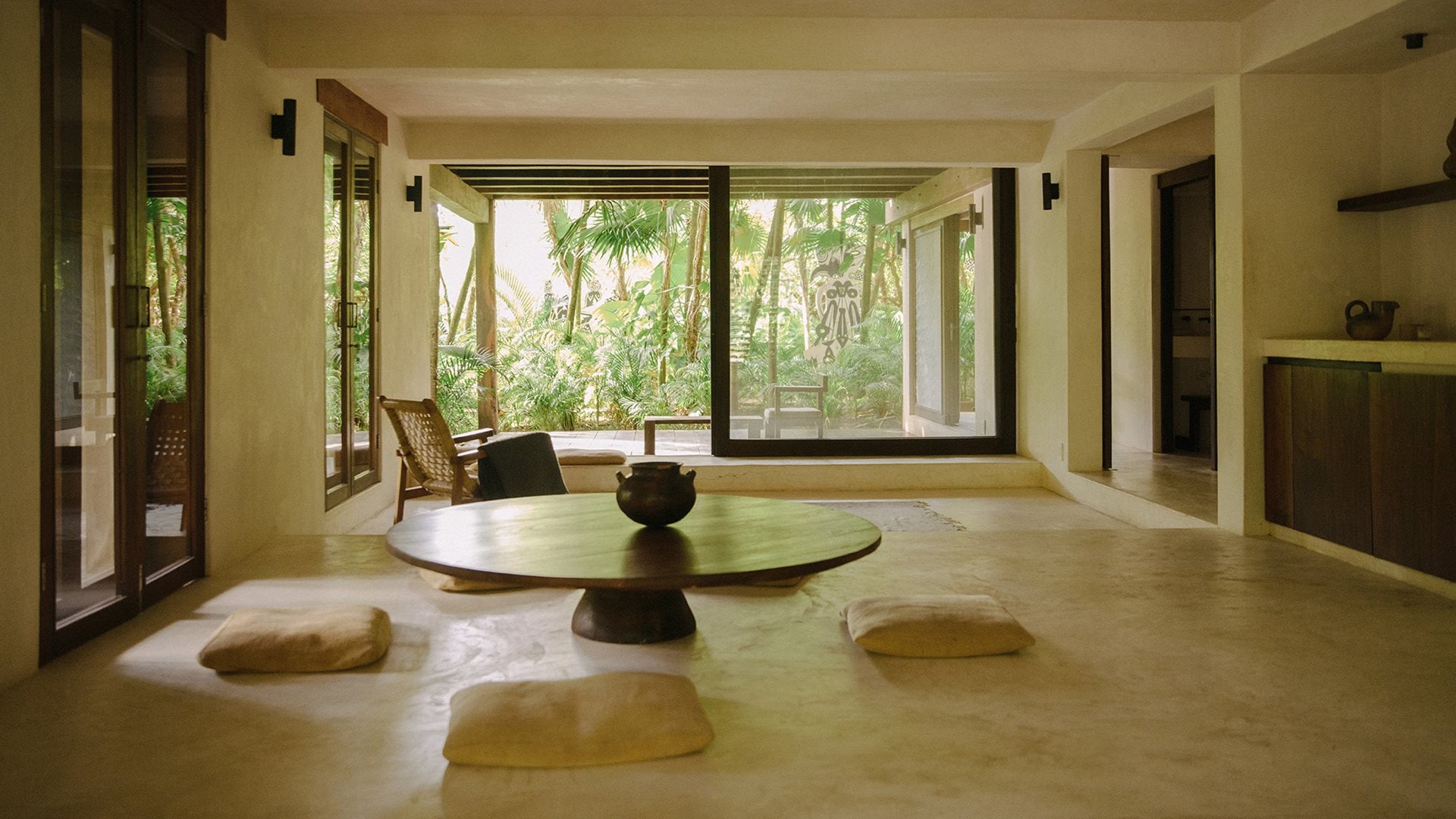 Tulum Treehouse, Mexico — Minimalist meditation room with circular table, floor cushions, tropical garden views through expansive windows, natural limestone flooring, warm neutral palette.