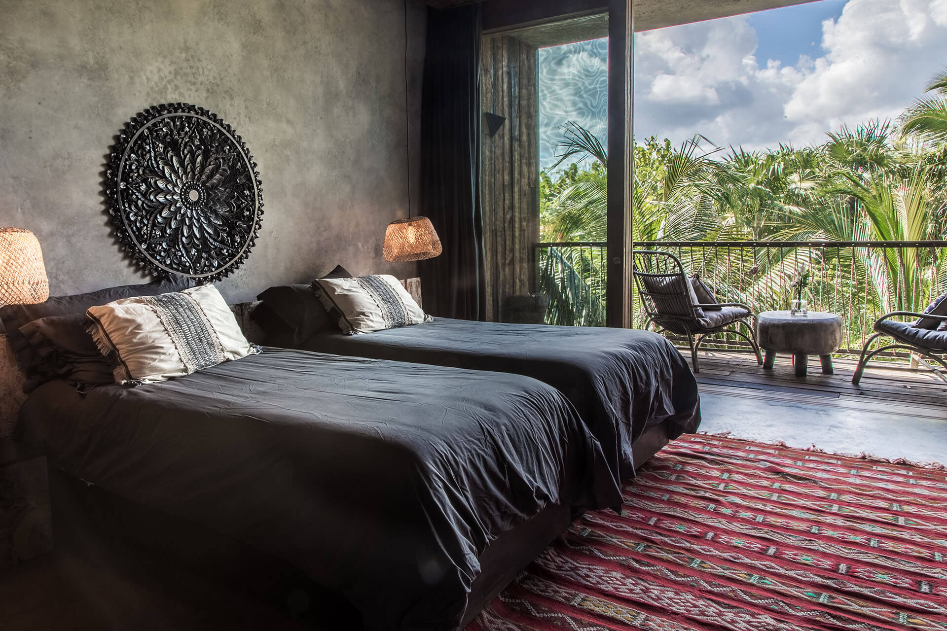 Be Tulum, Mexico — Luxury bedroom with dual beds, concrete walls, ornamental wall art, woven lamps, and ocean-view terrace overlooking tropical palms.