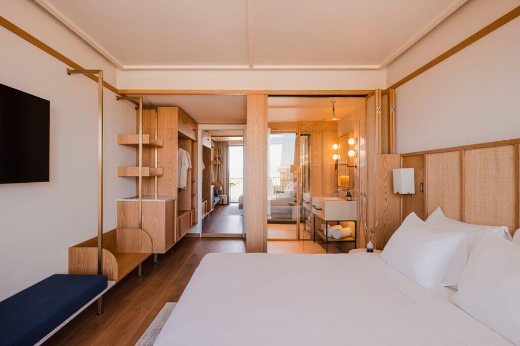 Aethos Ericeira, Portugal — Minimalist hotel suite with warm wood paneling, floor-to-ceiling wardrobe, cream bedding, and floor-to-ceiling windows overlooking coastal landscape.
