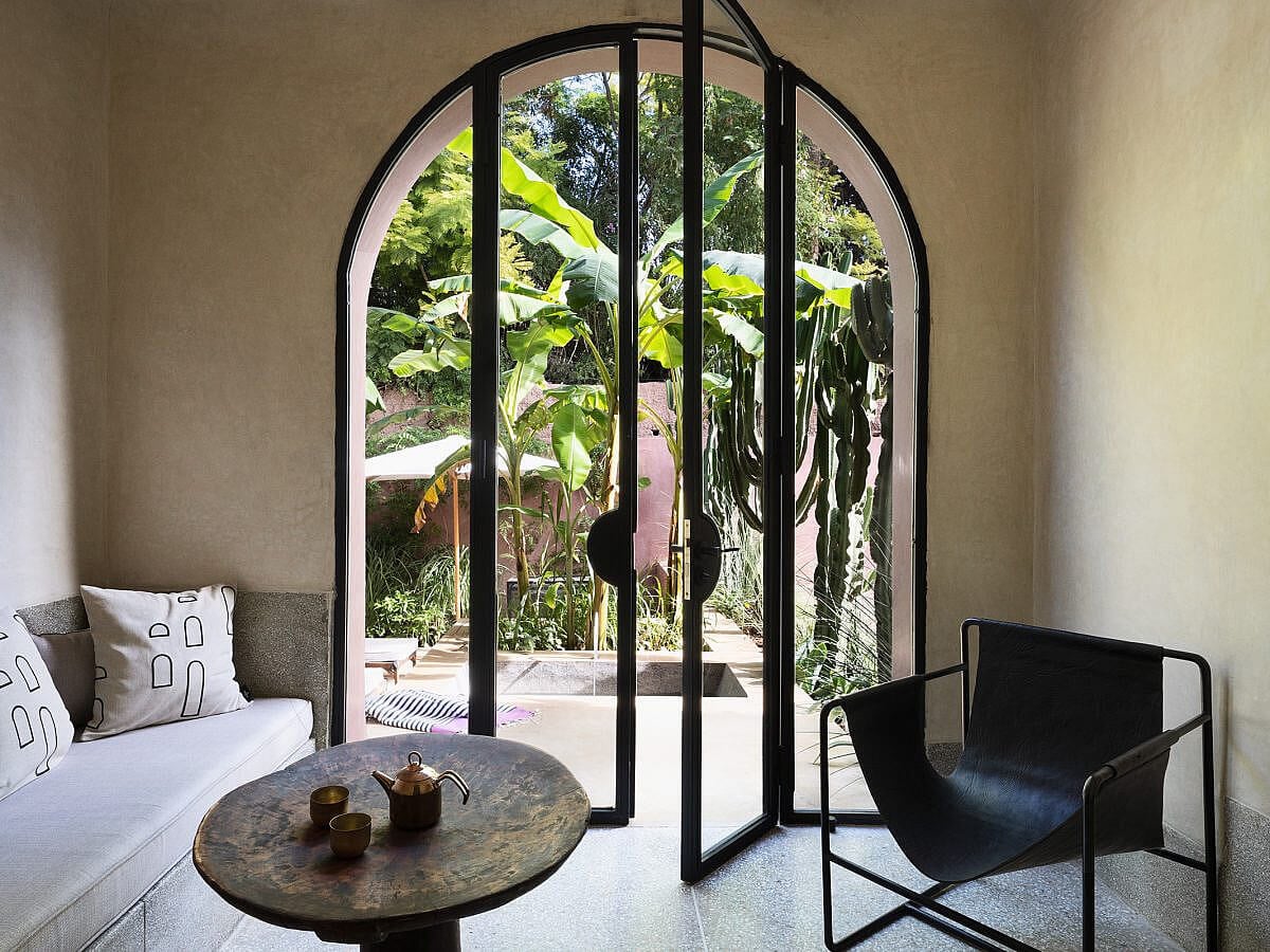Maison Brummell Majorelle, Africa — Arched black-framed glass doors open to lush tropical garden, with brass side table, white upholstered seating, and black leather chair.