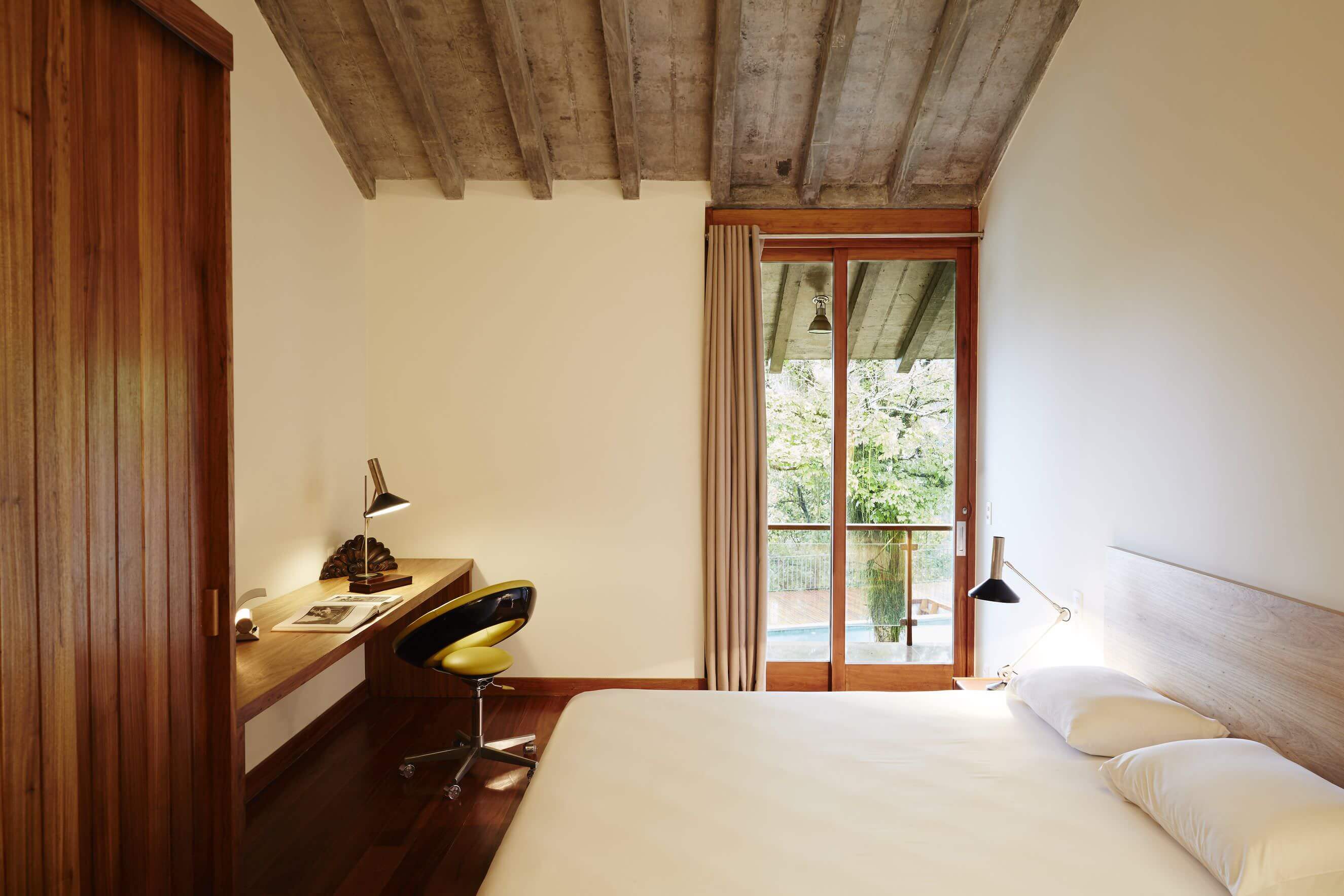 Chez Georges, South America — Minimalist bedroom with exposed timber ceiling beams, wooden desk, floor-to-ceiling glass doors opening to pool and garden views, warm natural light.