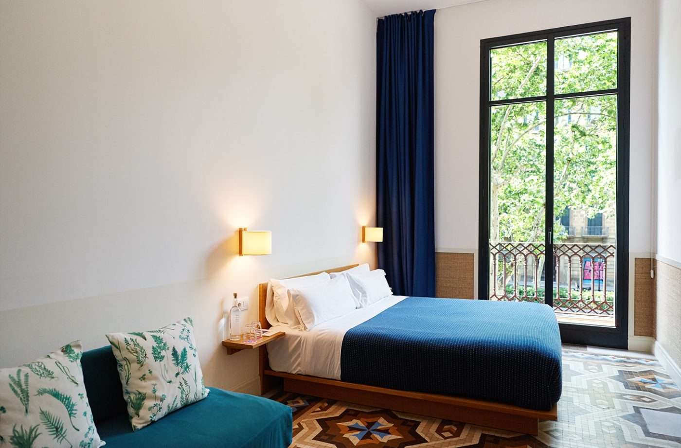 Casa Bonay, Europe — Minimalist bedroom with blue upholstered bed, forest-patterned pillow, brass wall sconces, navy curtains, parisian balcony doors overlooking ivy-covered facade.