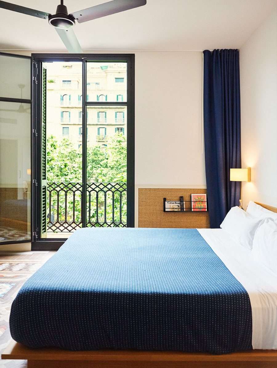 Casa Bonay, Barcelona, Spain — Modern bedroom with blue wool bedspread, black-framed French doors opening to balcony, dark navy curtains, ceiling fan, minimalist cork headboard, city views.