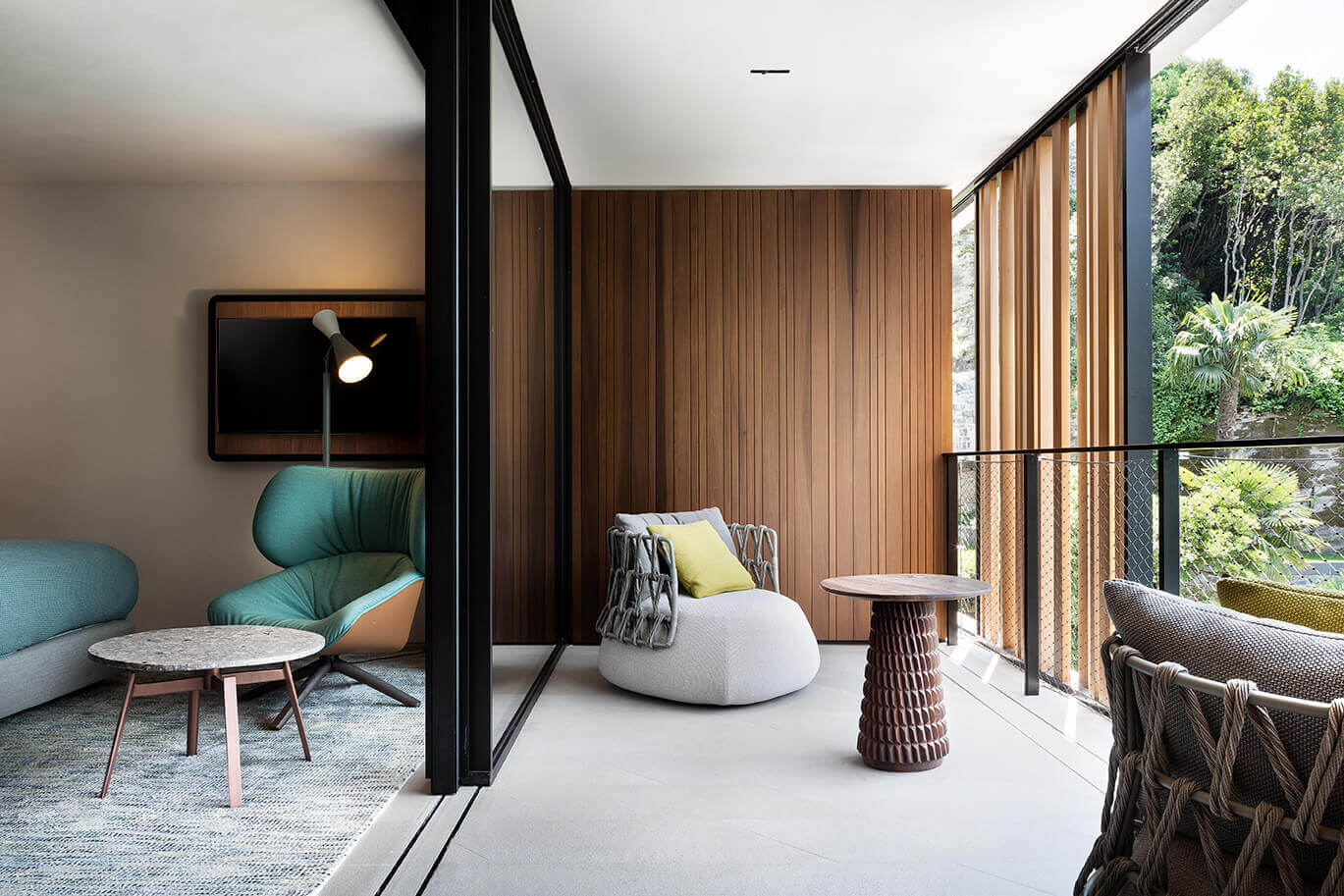 il Sereno Lago di Como, Italy — Modern lounge with teak wood paneling, turquoise curved chair, white bean bag seating, woven side tables, and floor-to-ceiling windows overlooking alpine forest.