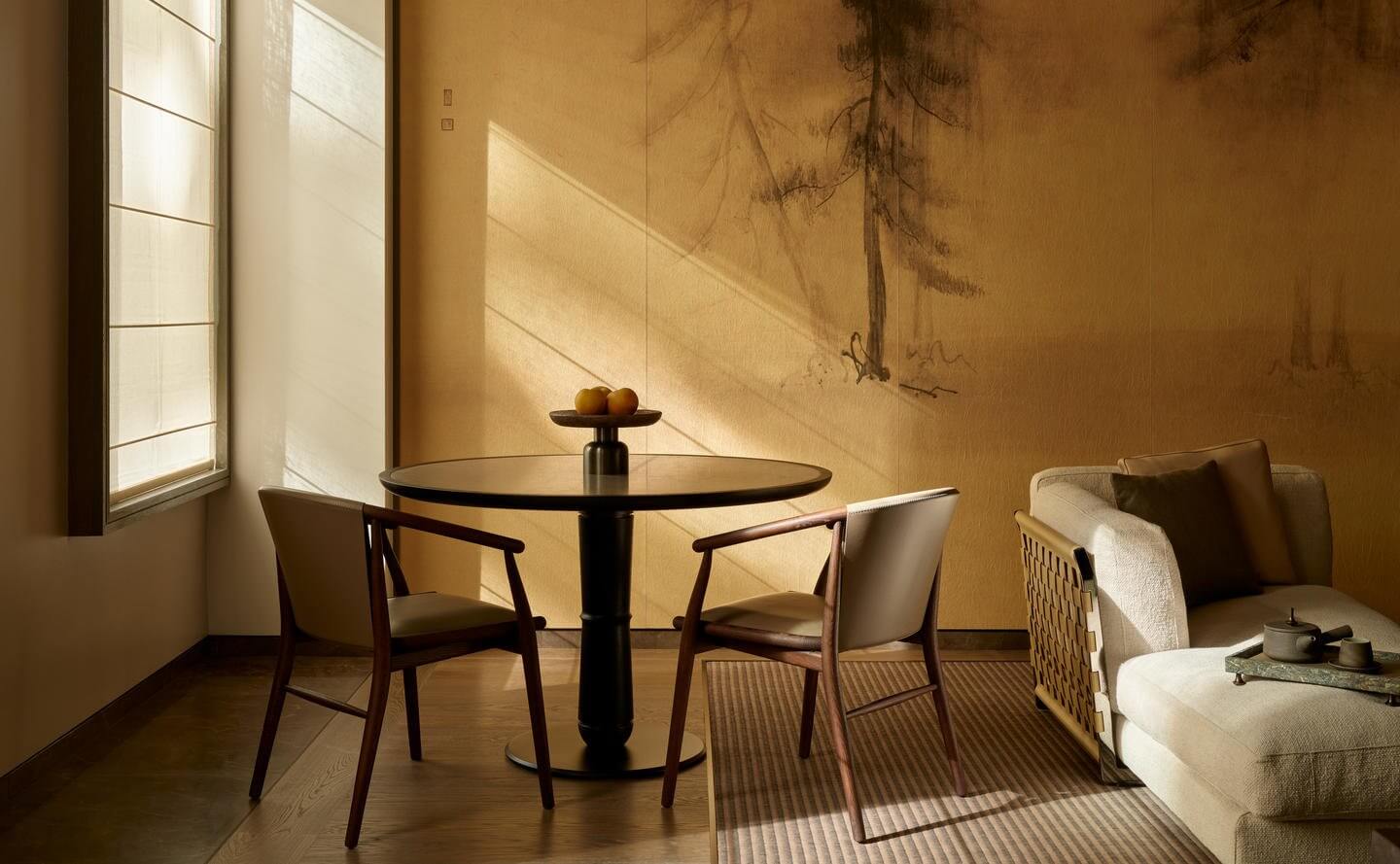Aman New York, North America — Minimalist dining room with round black table, mid-century chairs, ochre walls, and soft natural light casting tree shadows.