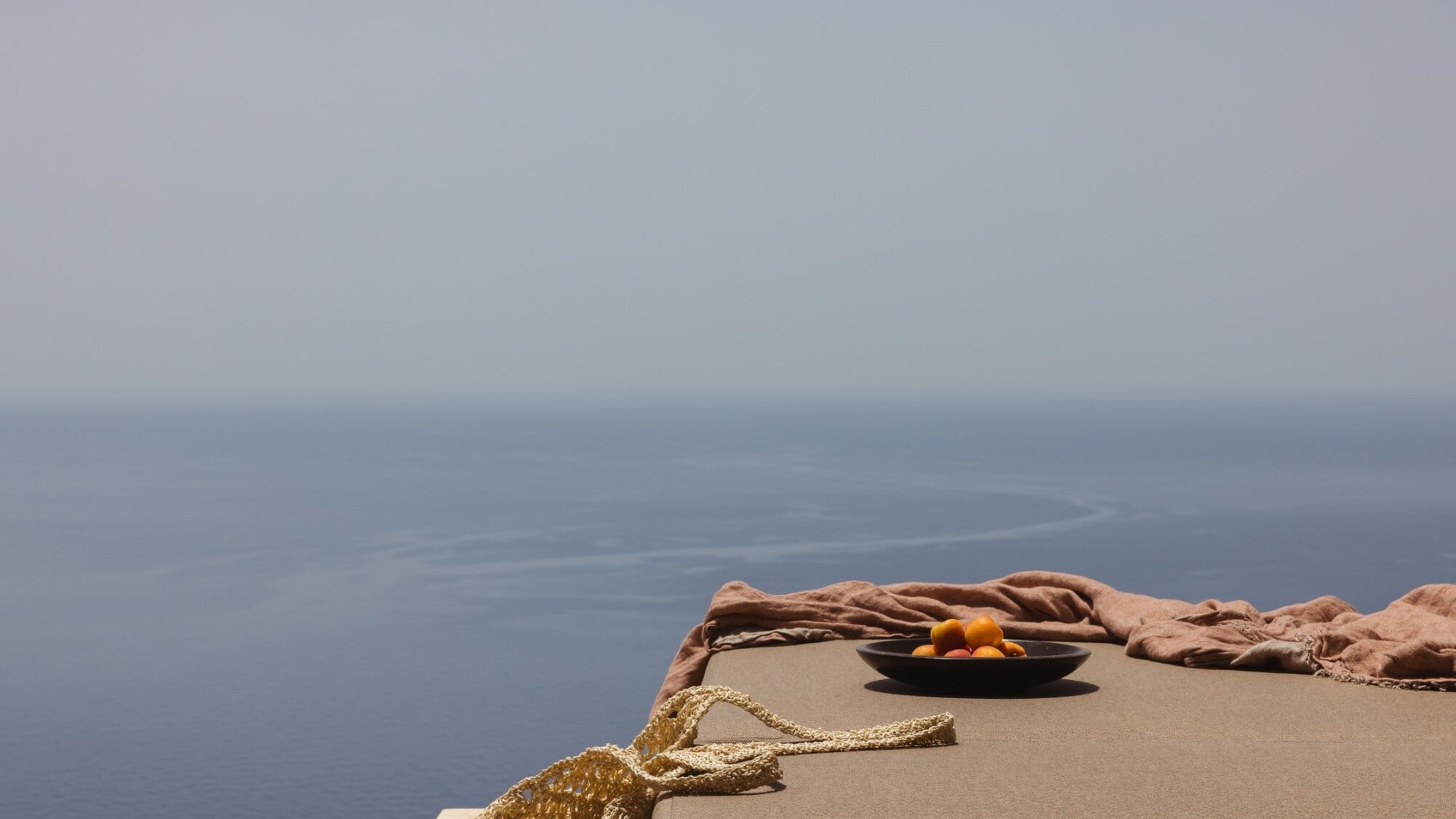 Gundari, Greece — Coastal terrace with linen cloth, golden rope, black ceramic bowl of citrus overlooking misty Aegean Sea at dawn.
