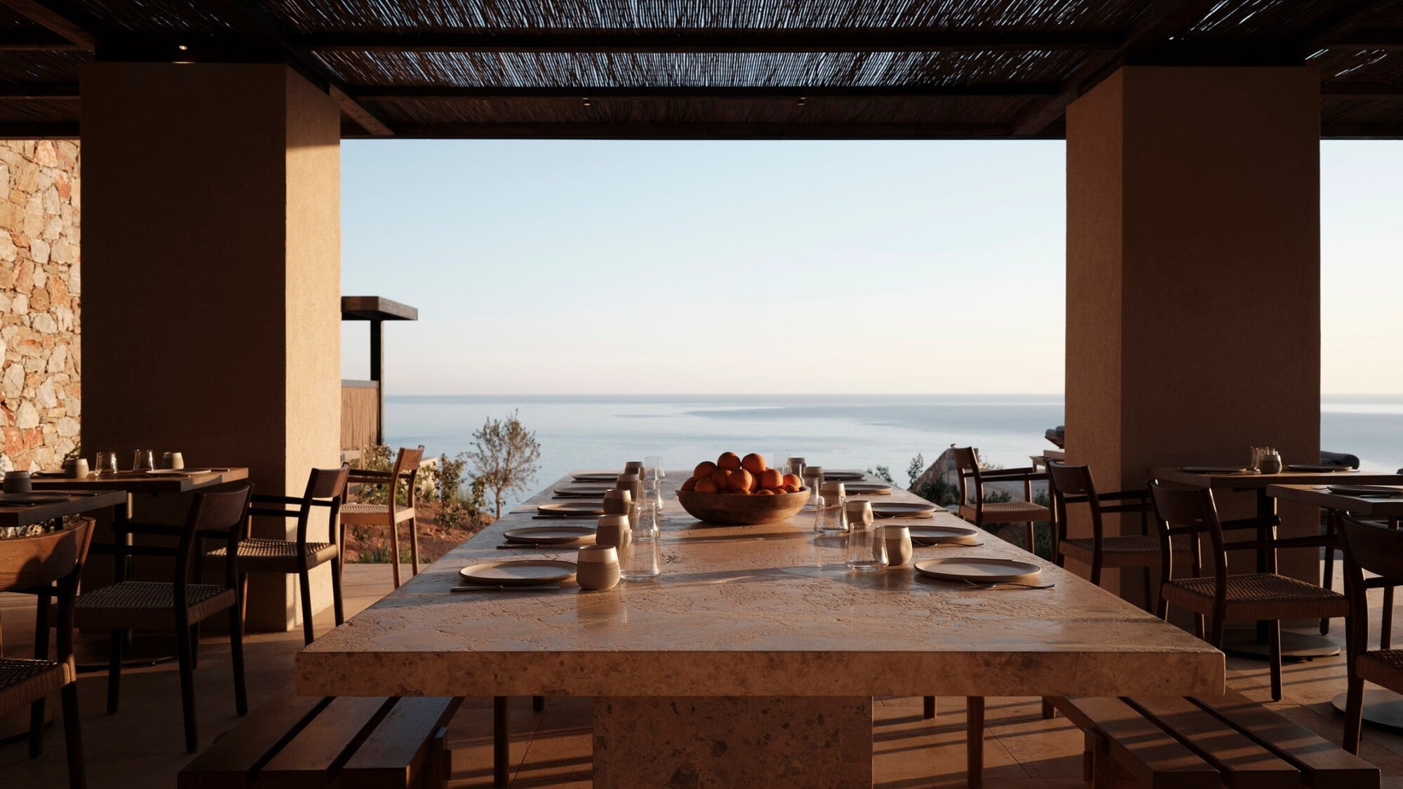 Gundari, Greece — Seaside dining pavilion with travertine table, linen chairs, and open-air terrace overlooking Aegean Sea at sunset.