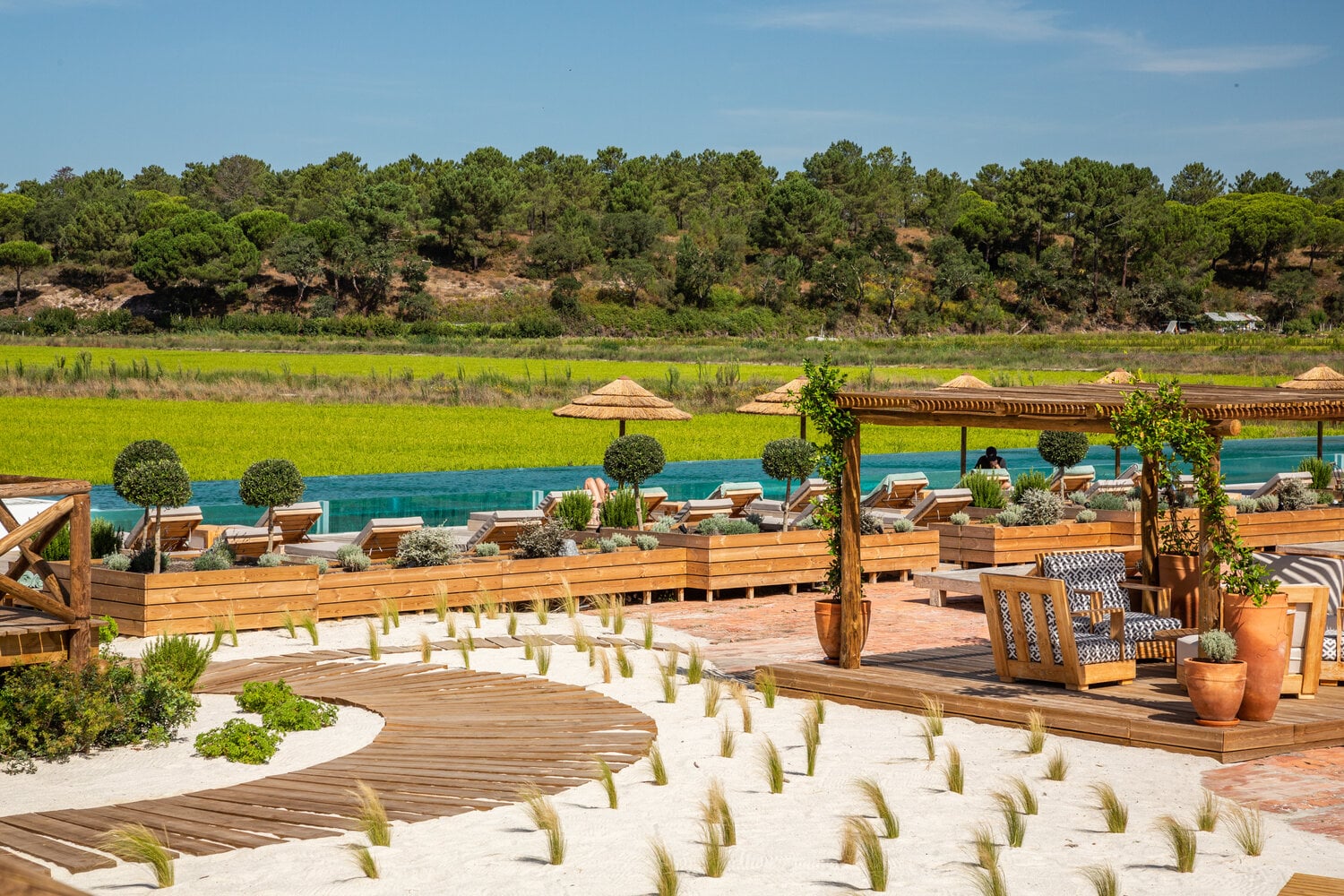 Quinta da Comporta, Portugal — Riverside beach club with teak loungers, woven umbrellas, wooden pergolas, and rice paddies beyond turquoise water.