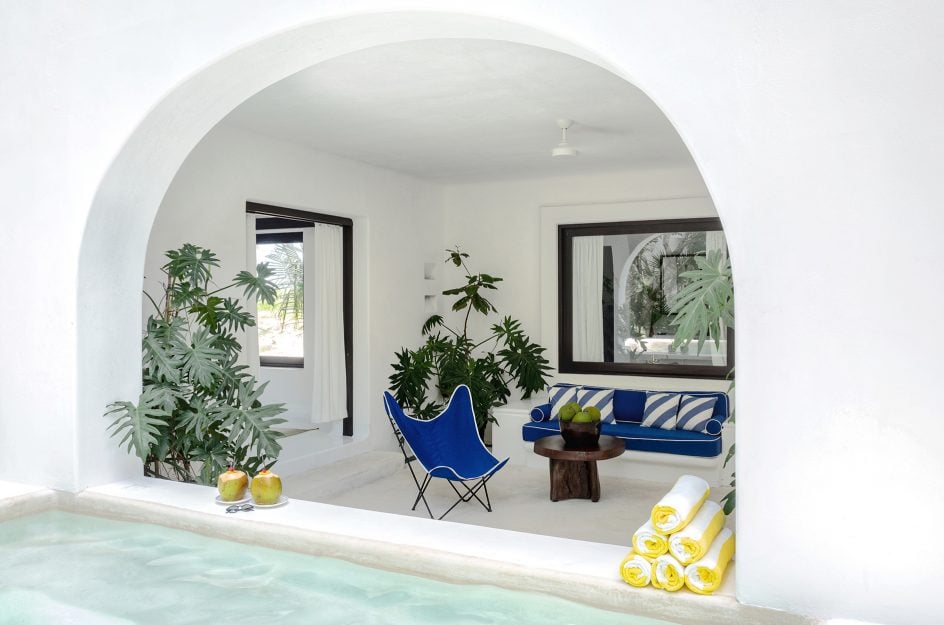 Hotel Esencia, Mexico — Whitewashed archway opening to Mediterranean-style loggia with blue butterfly chair, wooden side table, potted plants, and yellow rolled towels beside pool.