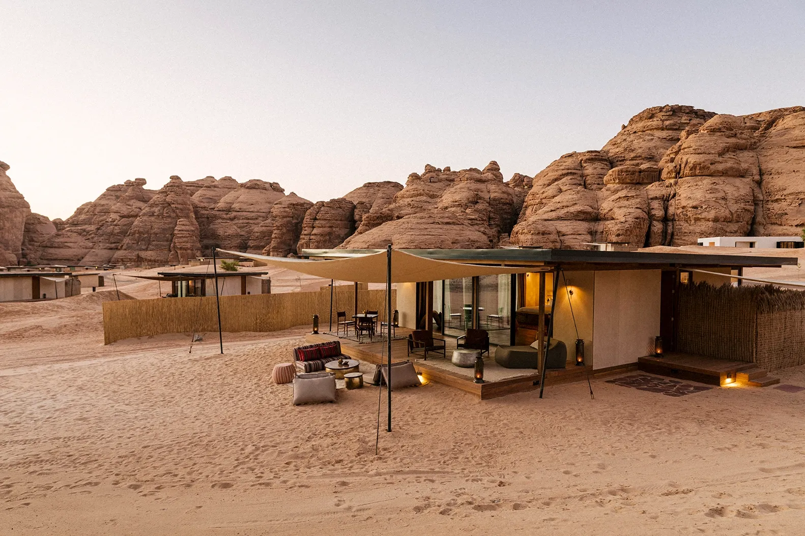 Habitas AlUla, Saudi Arabia — Contemporary pavilions with canvas canopies on sand beneath towering red sandstone formations at dusk, ambient lighting glowing warm.