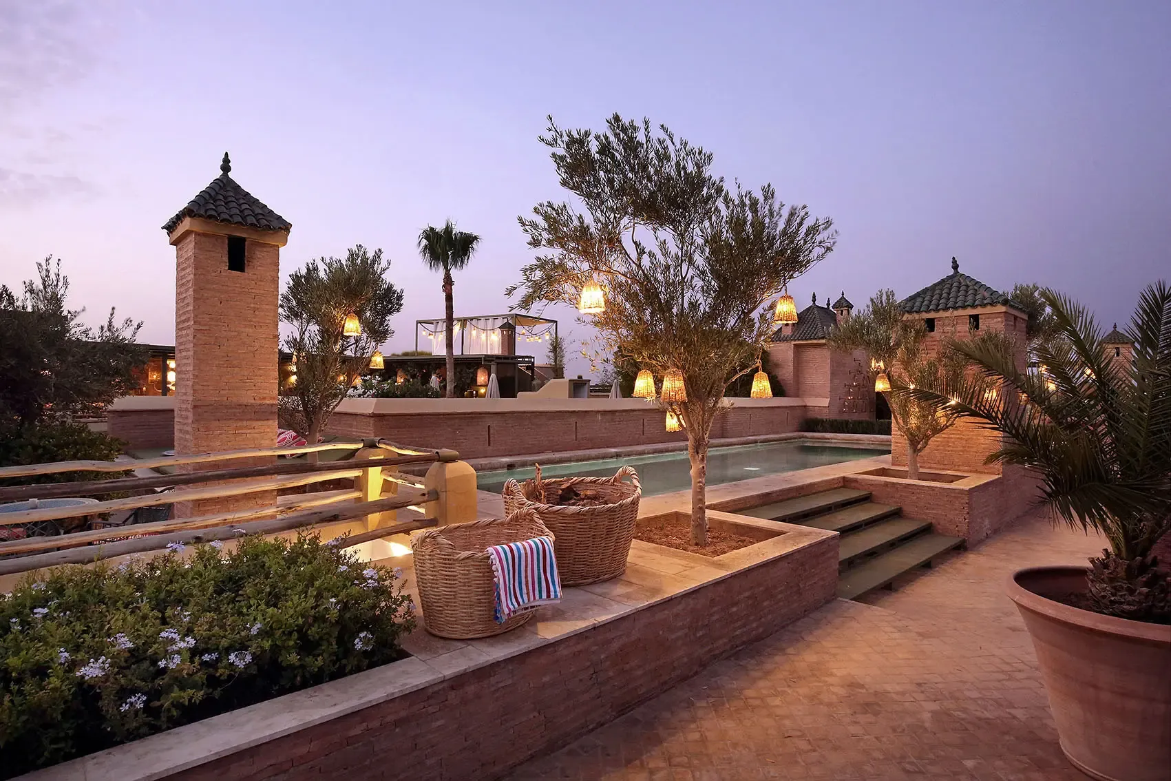 El Fenn, Morocco — Twilight terrace with riad architecture, illuminated brass lanterns, clay-tiled roofscape, palm trees, and reflecting pool at dusk.