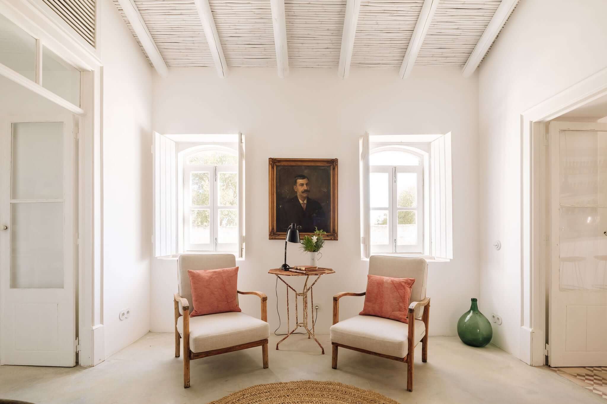 Hospedaria, Europe — Minimalist reception hall with symmetrical arched windows, wooden mid-century chairs, coral cushions, portrait painting, and natural light.