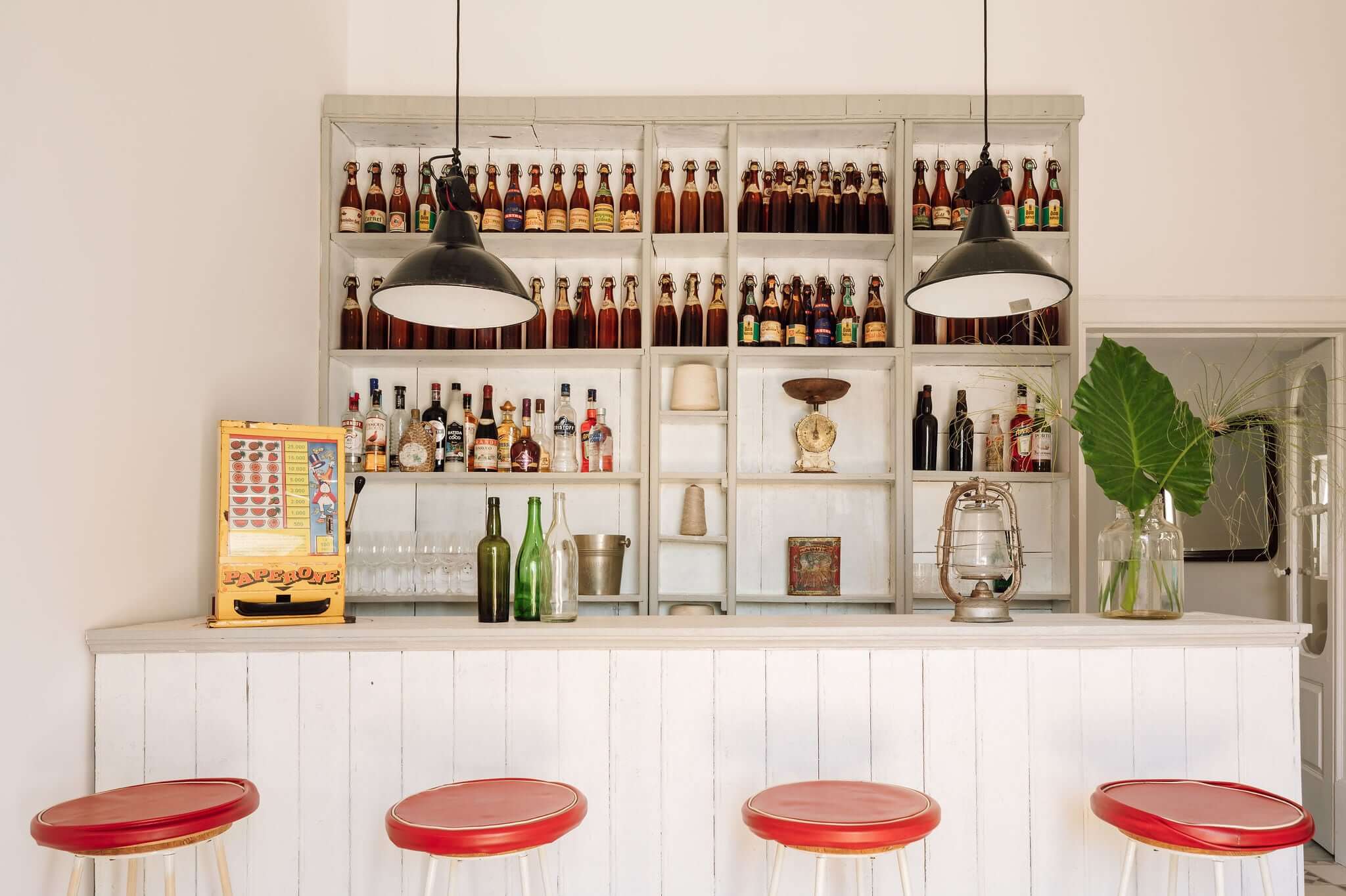 Hospedaria, Europe — Modern bar interior with white shelving, vintage beer bottles, black pendant lights, red vinyl stools, and curated decorative objects.