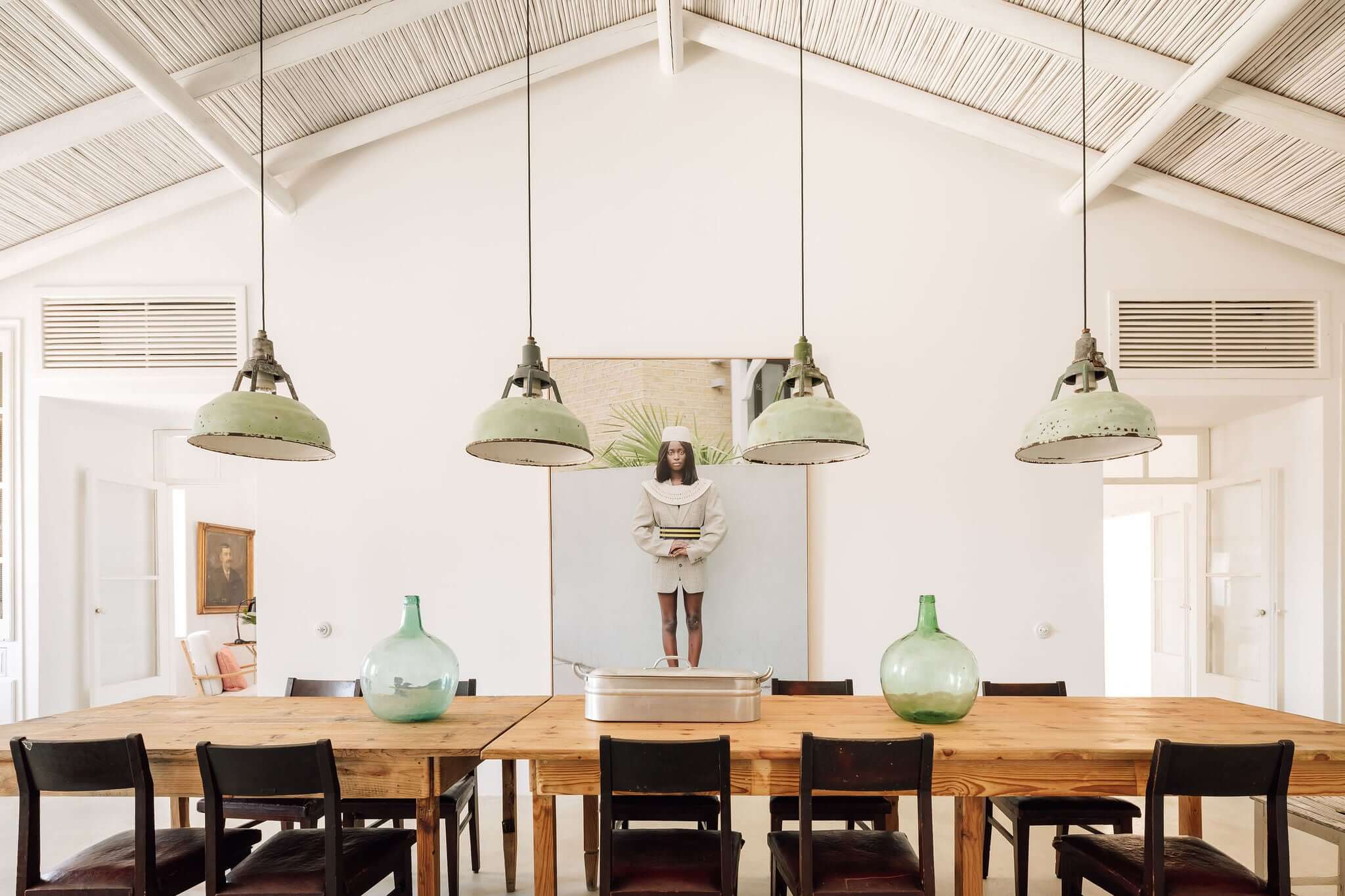 Hospedaria, Europe — Dining room with vaulted ceiling, sage pendant lights, wooden table, black chairs, green glass vessels, and gallery wall with portrait.