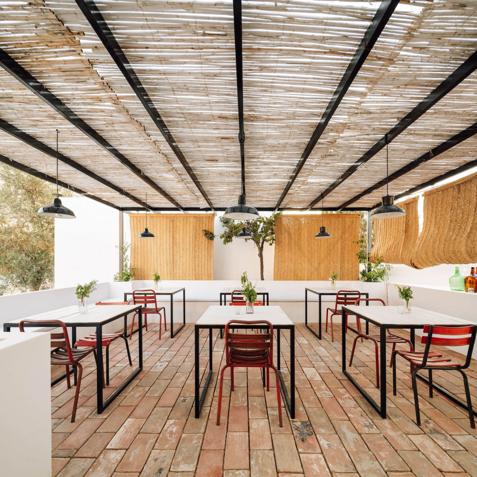 Hospedaria, Europe — Roofed terrace with woven reed ceiling, terracotta tile flooring, red chairs, black tables, and pendant lights under natural daylight.