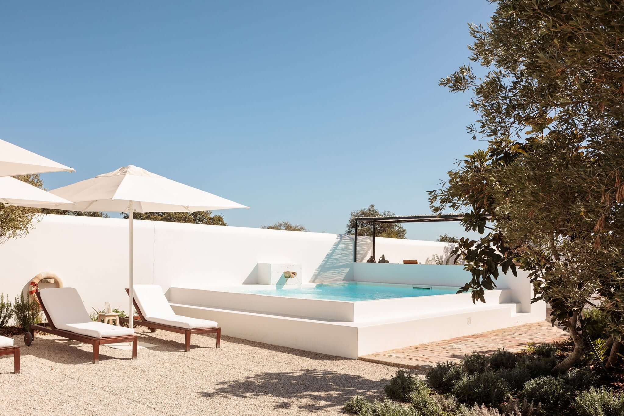Hospedaria, Europe — Minimalist poolside terrace with turquoise water basin, white sculptural architecture, wooden loungers, umbrella, and olive trees under clear sky.