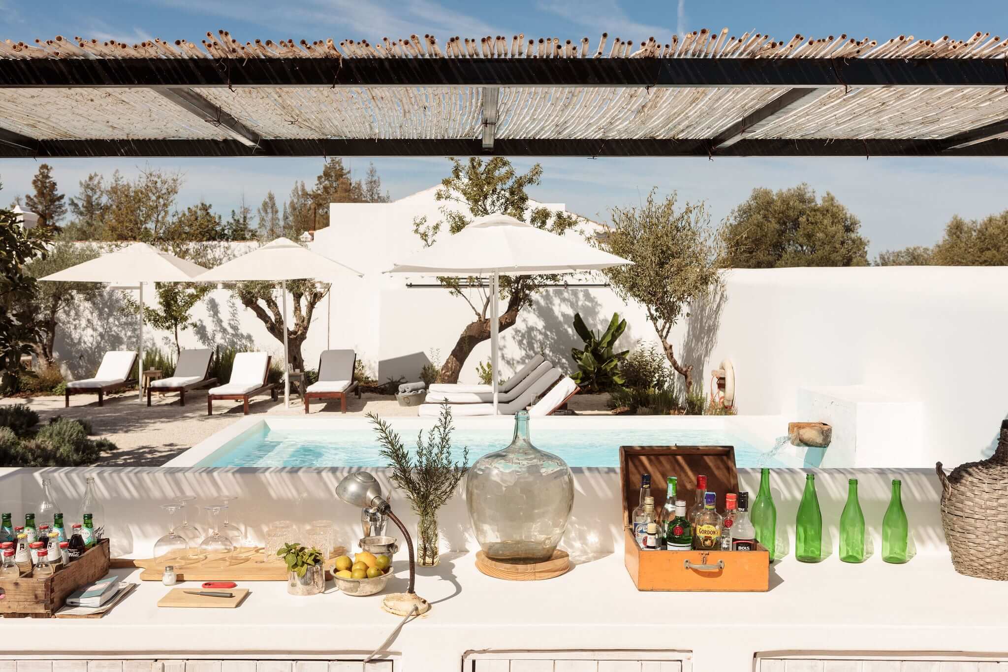 Hospedaria, Europe — Whitewashed courtyard with pool, olive trees, lounge chairs beneath umbrellas, wooden pergola, and bar setup under Mediterranean light.