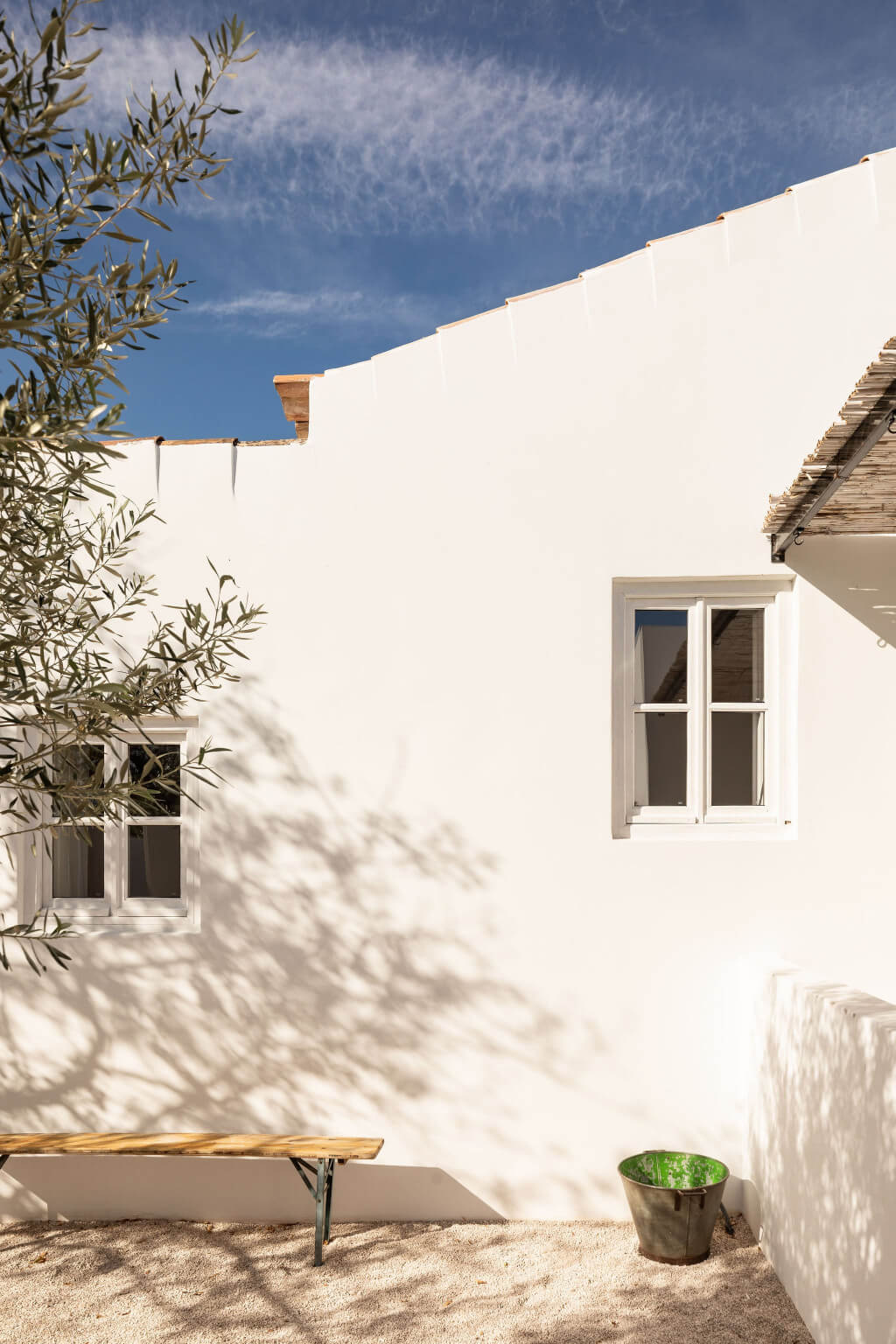 Hospedaria, Europe — Whitewashed courtyard with olive tree casting dappled shadows, modernist windows, wooden bench, potted plant, clear blue sky.