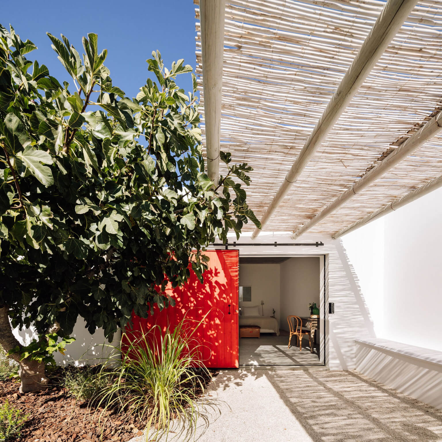 Hospedaria, Europe — Modern courtyard with slatted pergola casting geometric shadows, red accent wall, lush vegetation, and open glass doors to minimalist interior.