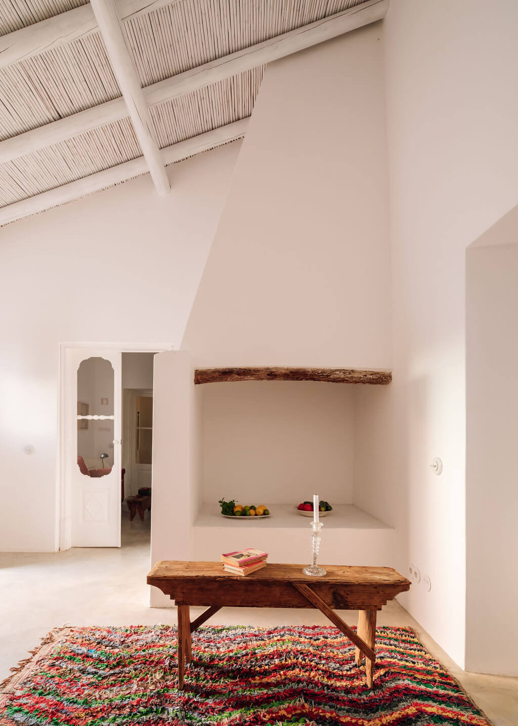 Hospedaria, Europe — Minimalist whitewashed room with wooden bench, colorful woven rug, recessed niche, and exposed beam ceiling detail.