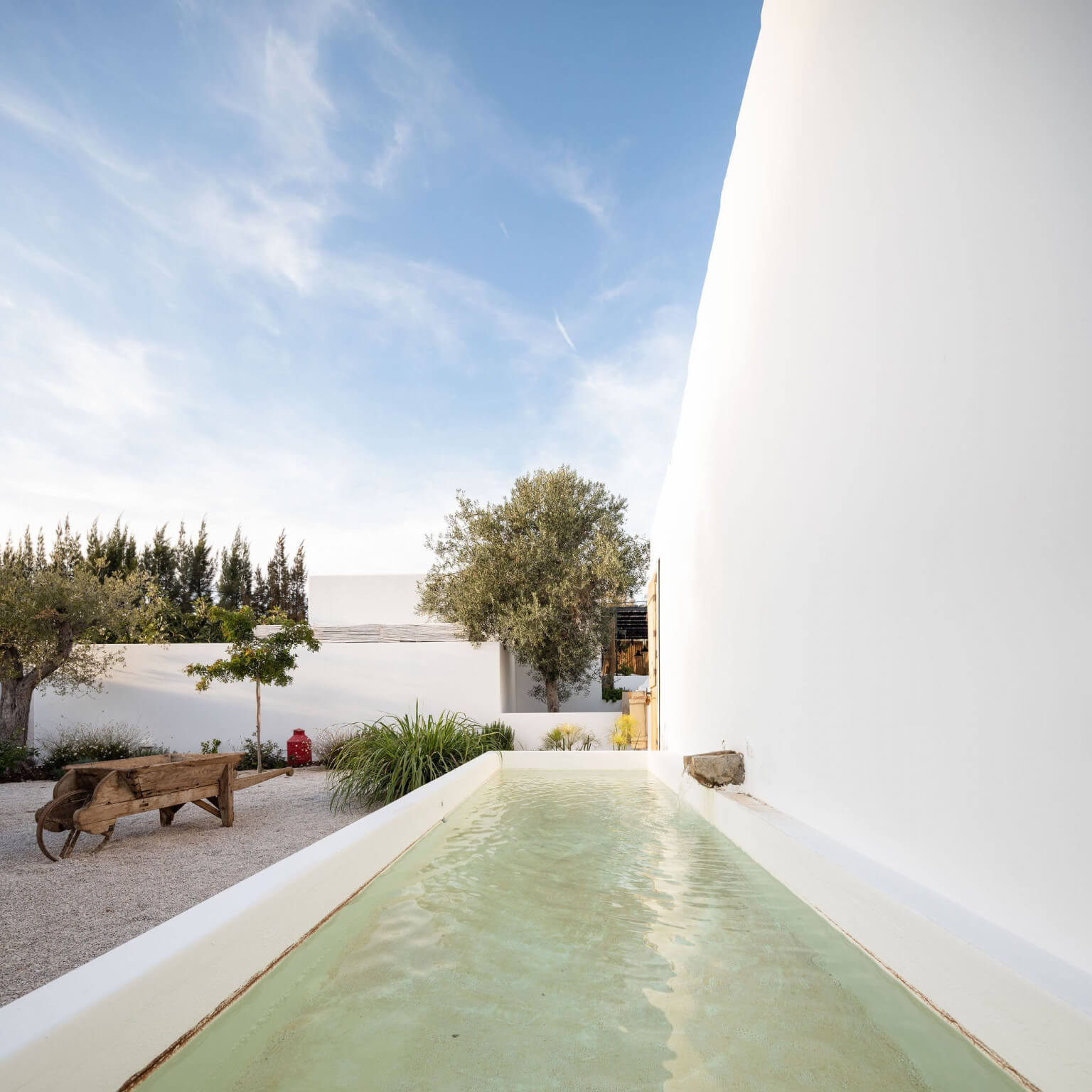 Hospedaria, Europe — narrow lap pool between white-walled structures with gravel courtyard, olive trees, and cypress beyond under clear sky.