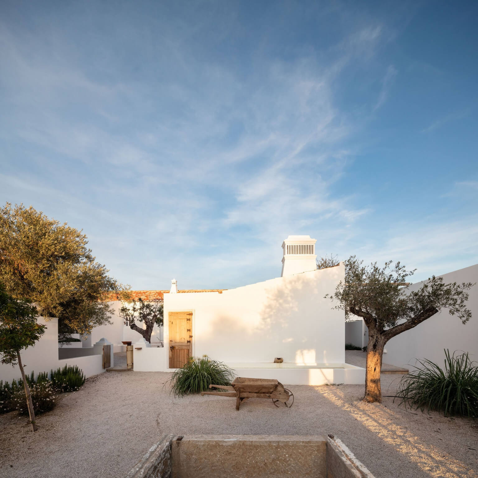 Hospedaria, Europe — Whitewashed courtyard with olive trees, wooden bench, and minimalist architecture under clear sky.
