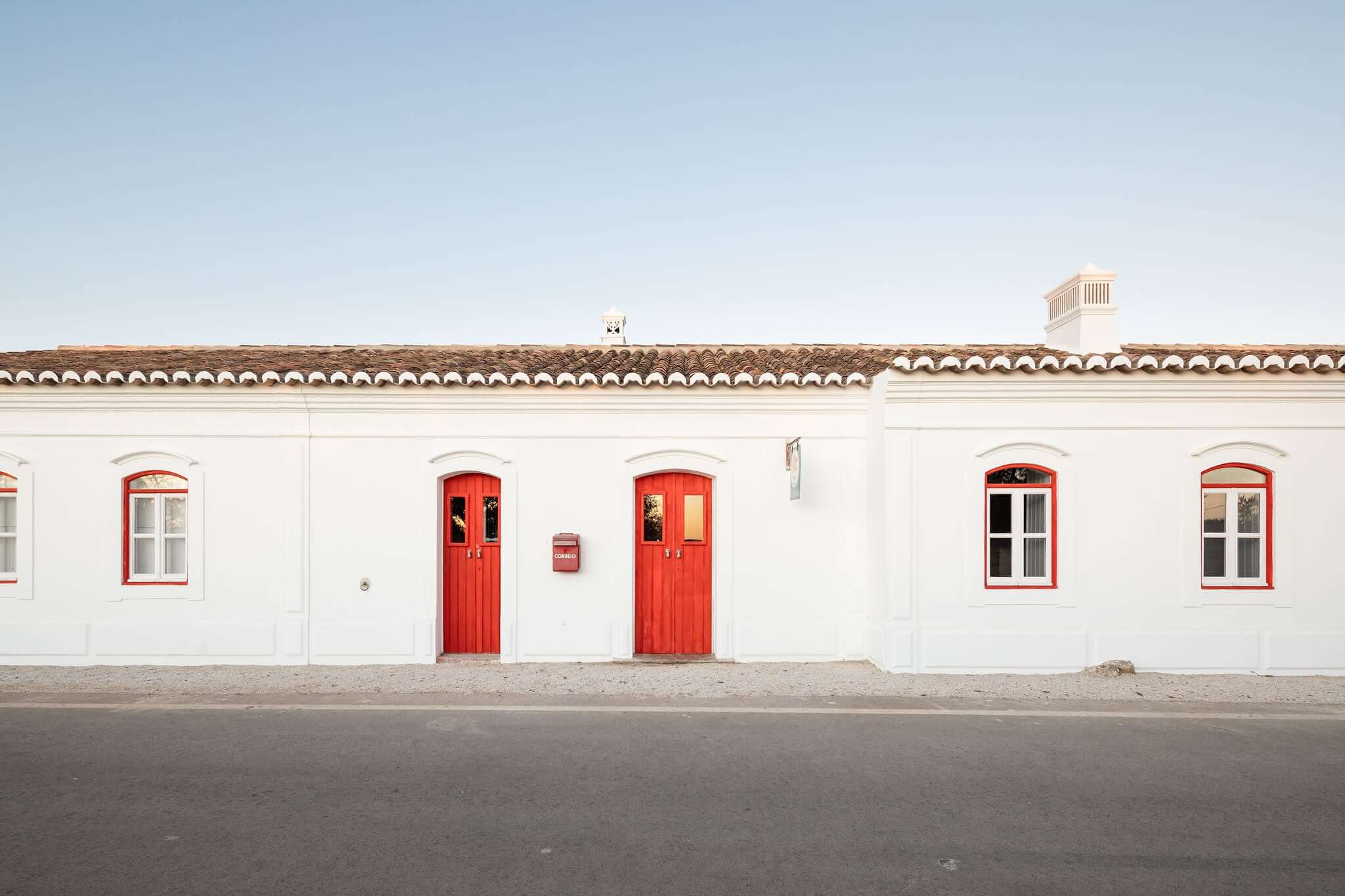Hospedaria, Europe — White-walled colonial building with terracotta roof, bright red doors and window frames, minimalist facade, clear blue sky.
