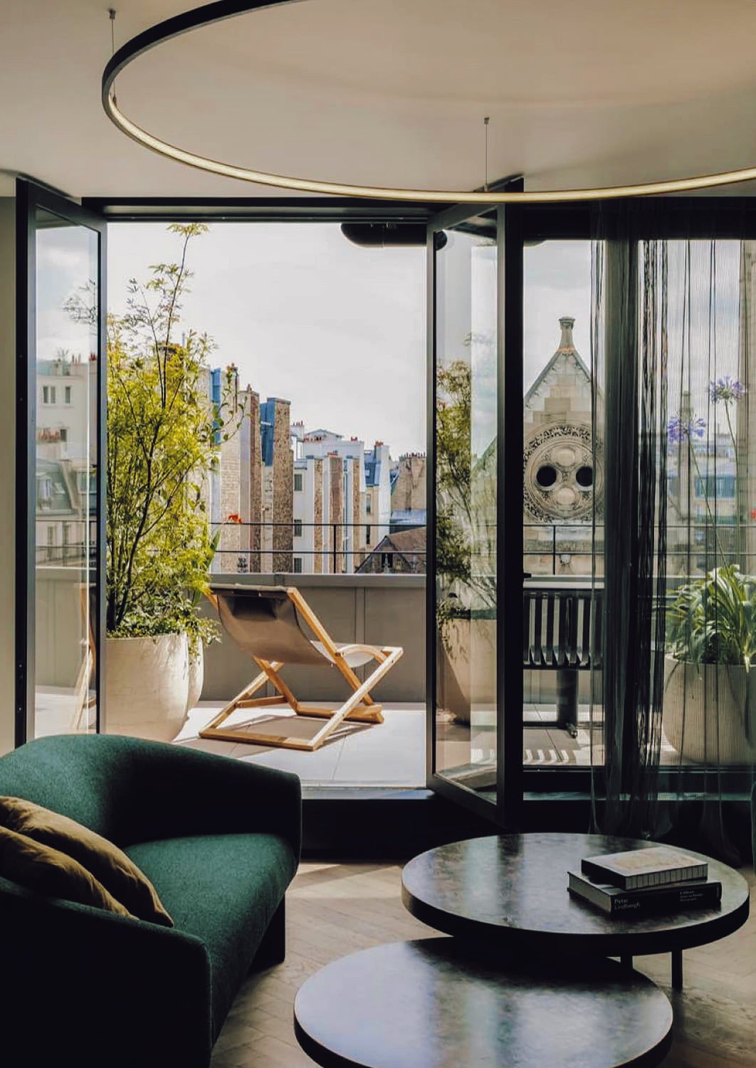 Hôtel National Des Arts, Paris, France — Modern living room with green curved chair, marble tables, and floor-to-ceiling black-framed doors opening to Parisian rooftop terrace with planted greenery.