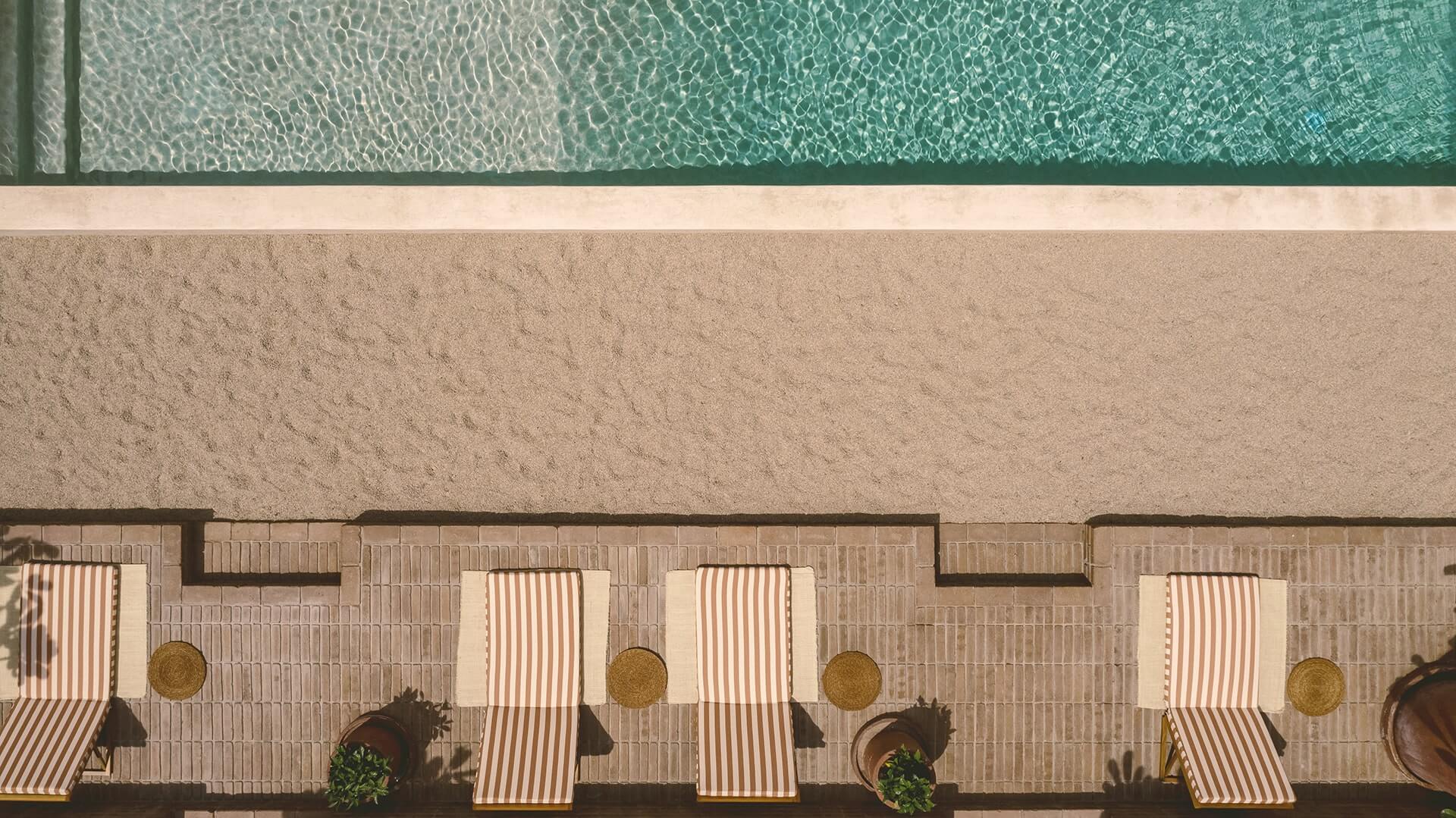Baja Club, Mexico — Aerial view of striped loungers and umbrellas on terracotta poolside deck beside turquoise water.