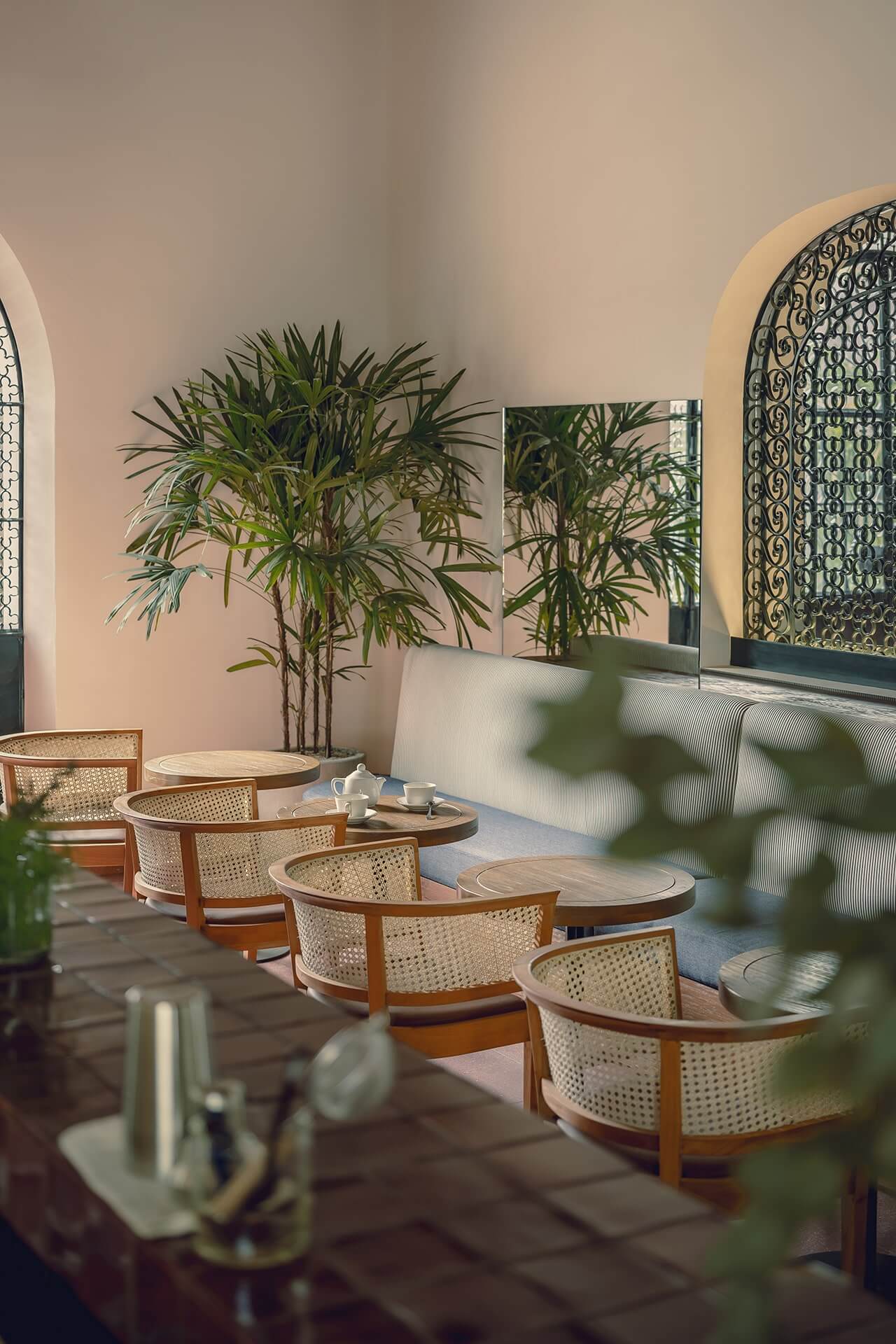 Baja Club Restaurant, Mexico — Woven rattan chairs with wooden frames arranged around pale stone tables in a sun-lit courtyard with arched alcoves, potted bamboo, and decorative latticed windows.