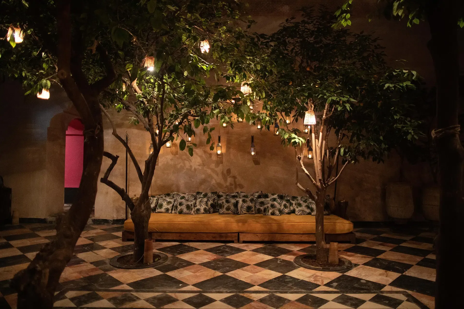 El Fenn, Marrakech, Morocco — Courtyard with lantern-lit trees, geometric tile flooring, cushioned bench against ochre wall, nighttime ambiance.