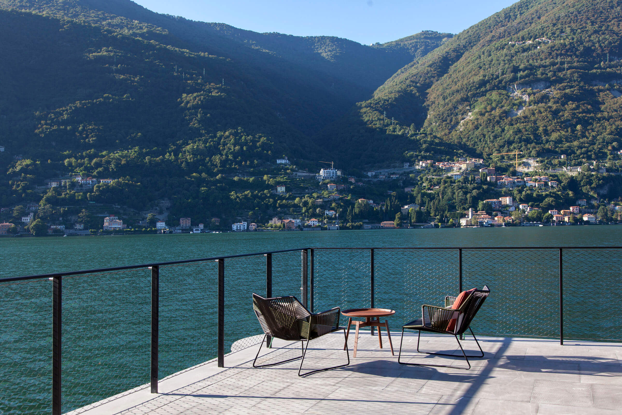 il Sereno Lago di Como, Europe — Woven chairs and small table on lakeside terrace with turquoise water, forested mountains, and lakeside village beyond.