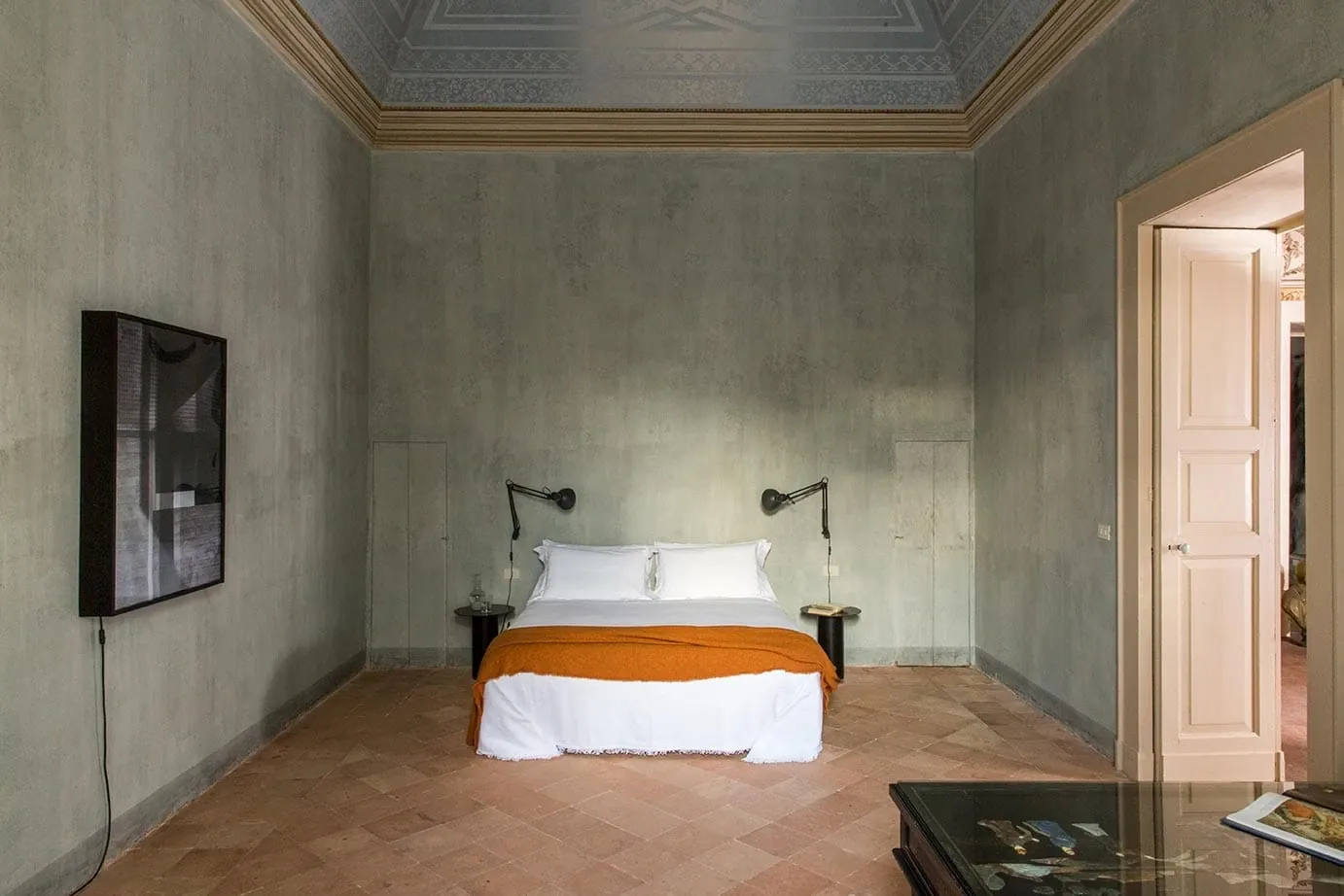 Palazzo Daniele, Europe — Minimalist bedroom with terracotta floors, sage plaster walls, iron sconces, and ornate coffered ceiling in luxury Italian palazzo hotel.