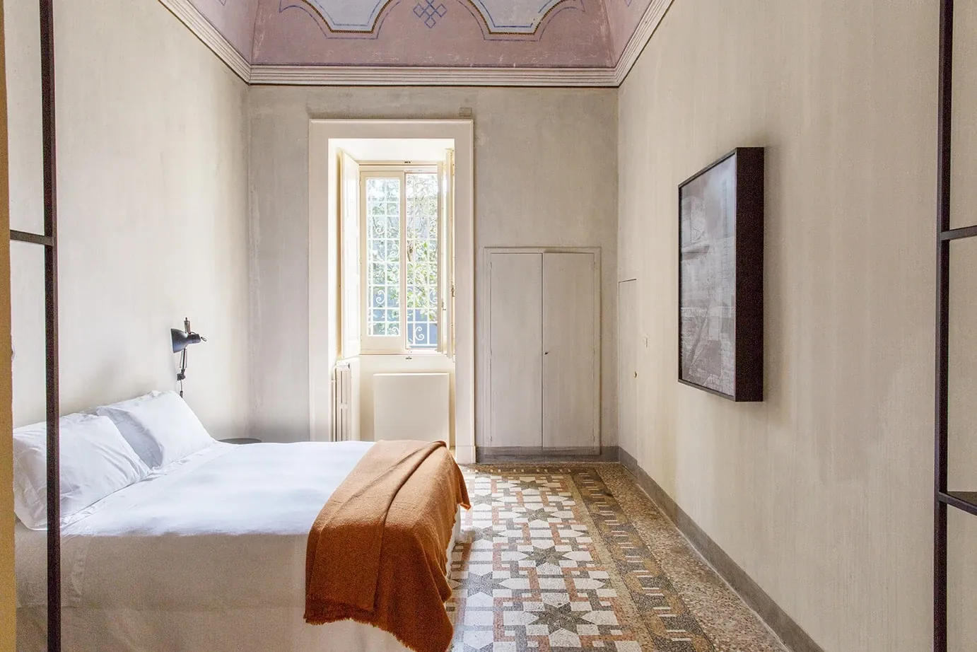 Palazzo Daniele, Europe — Minimalist bedroom with canopy bed, terracotta throw, geometric floor tiles, arched ceiling detail, and tall shuttered window overlooking gardens.