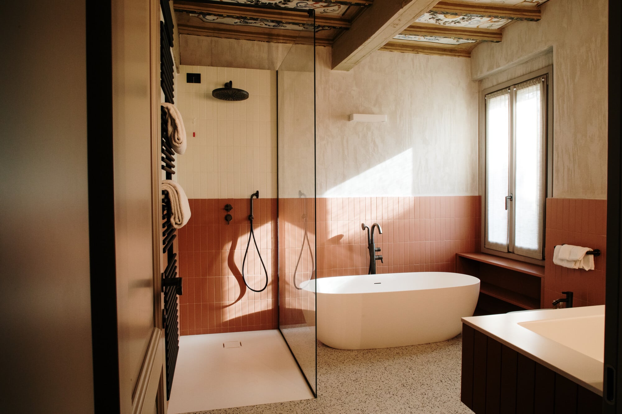 Roncolo 1888, Europe — Minimalist bathroom with terracotta tile accent wall, freestanding white oval bathtub, glass-enclosed shower, wooden ceiling beams, natural light.