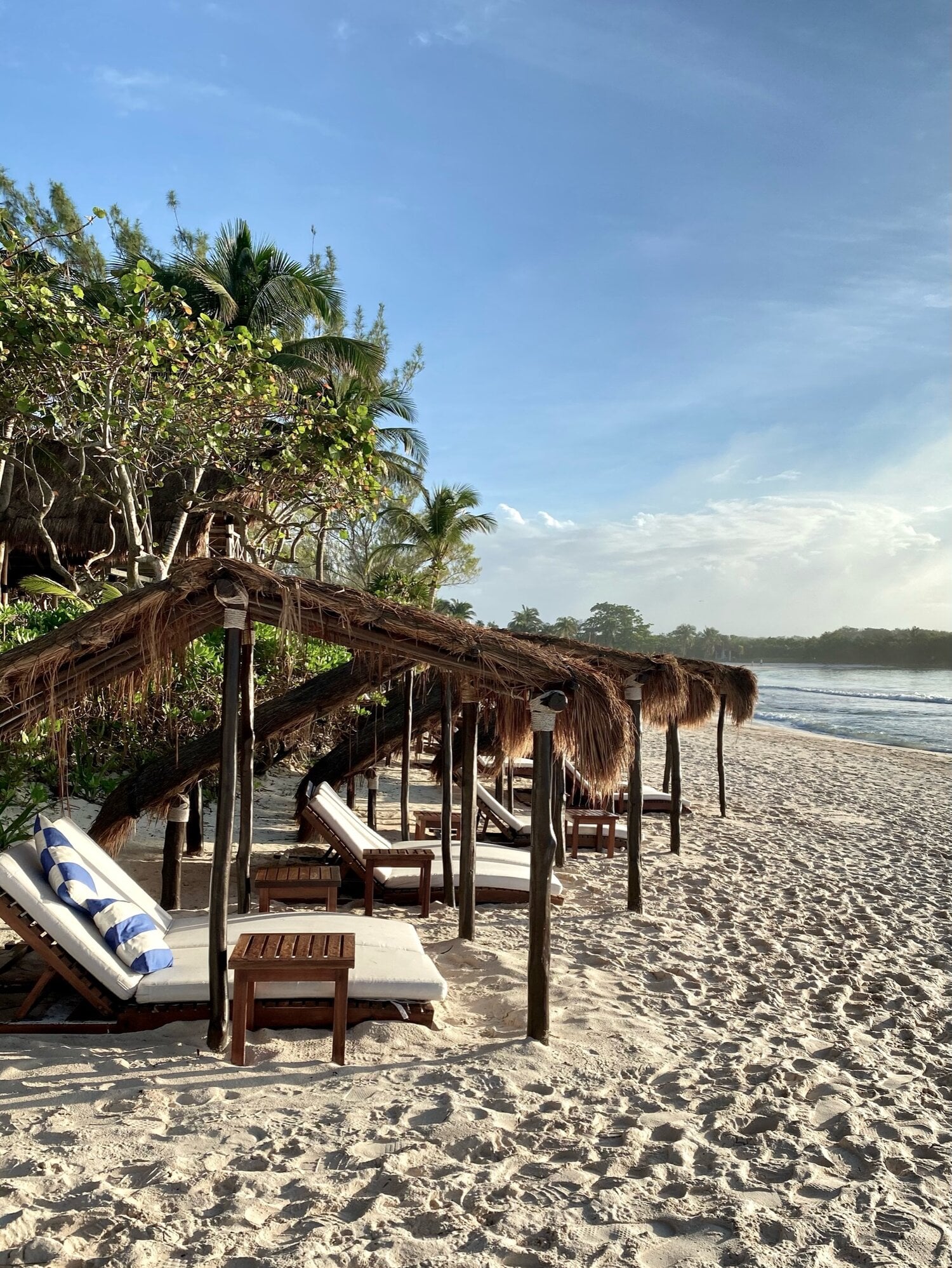Hotel Esencia, Mexico — Thatched beachfront cabanas with wooden loungers overlooking the Caribbean Sea, palm trees and sand.
