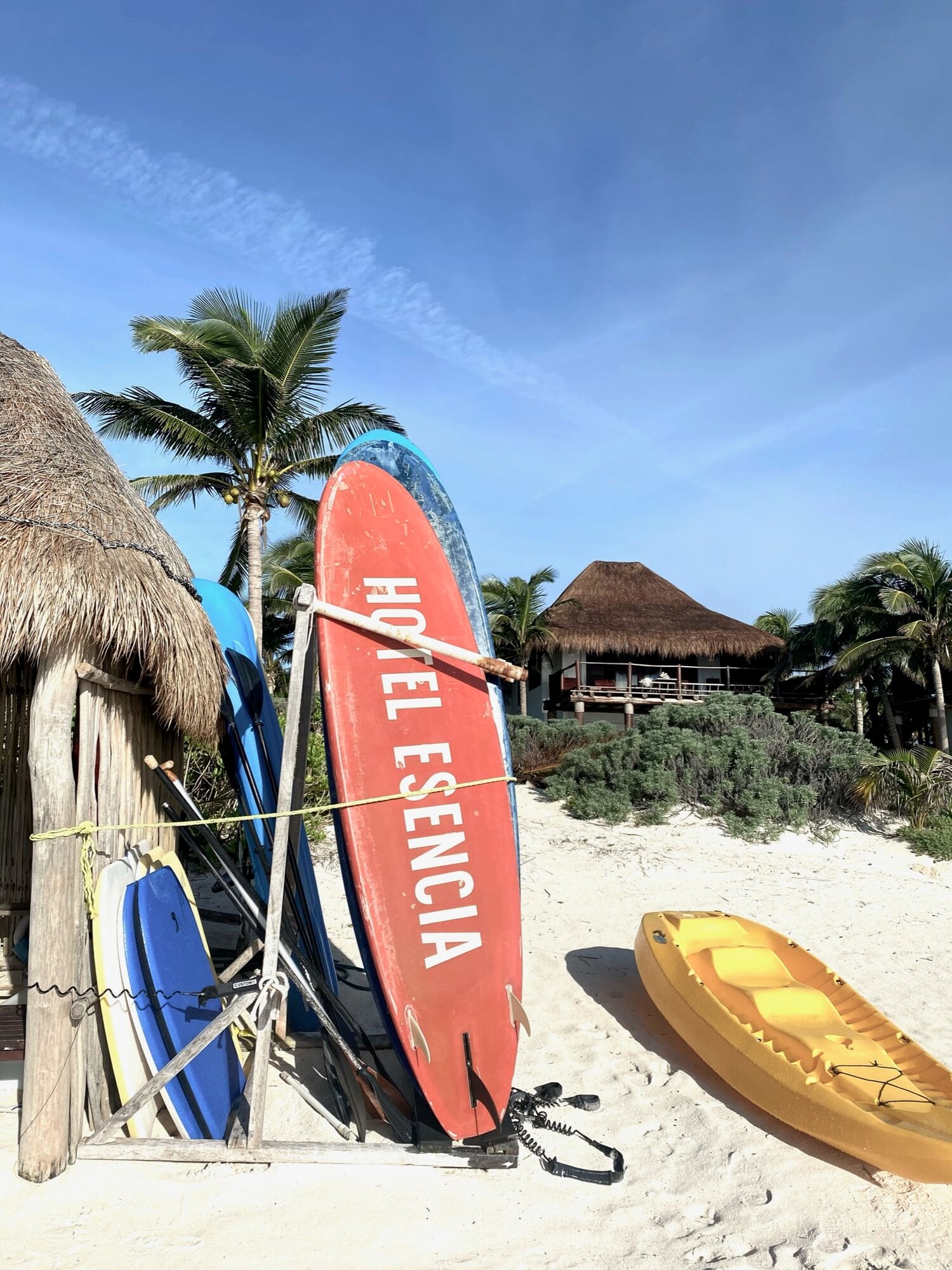 Hotel Esencia, Mexico — Red surfboard and yellow kayak on sand beach with thatched-roof bungalows and palm trees under clear sky.
