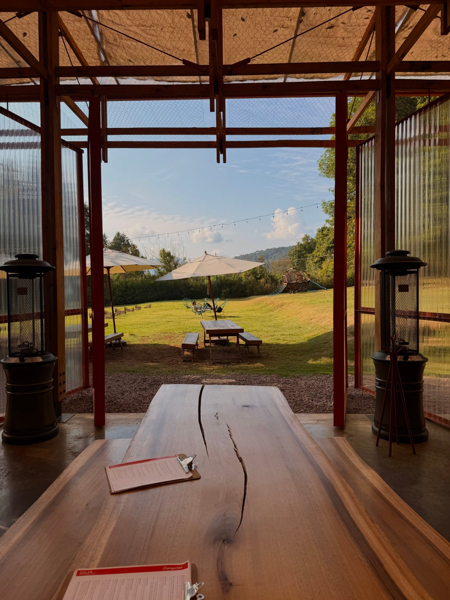 Solar, Mexico — Live-edge wood table in open-air pavilion overlooking manicured lawn, umbrellas, and forested hills at sunset.