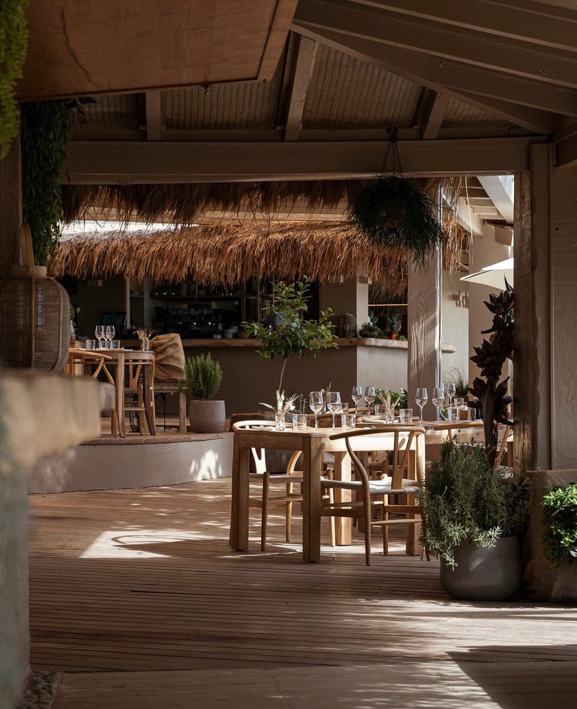 El Chiringuito, Balearic Islands, Spain — Thatched-roof beachside restaurant with wooden dining tables, natural light, potted plants, and minimalist Mediterranean design.