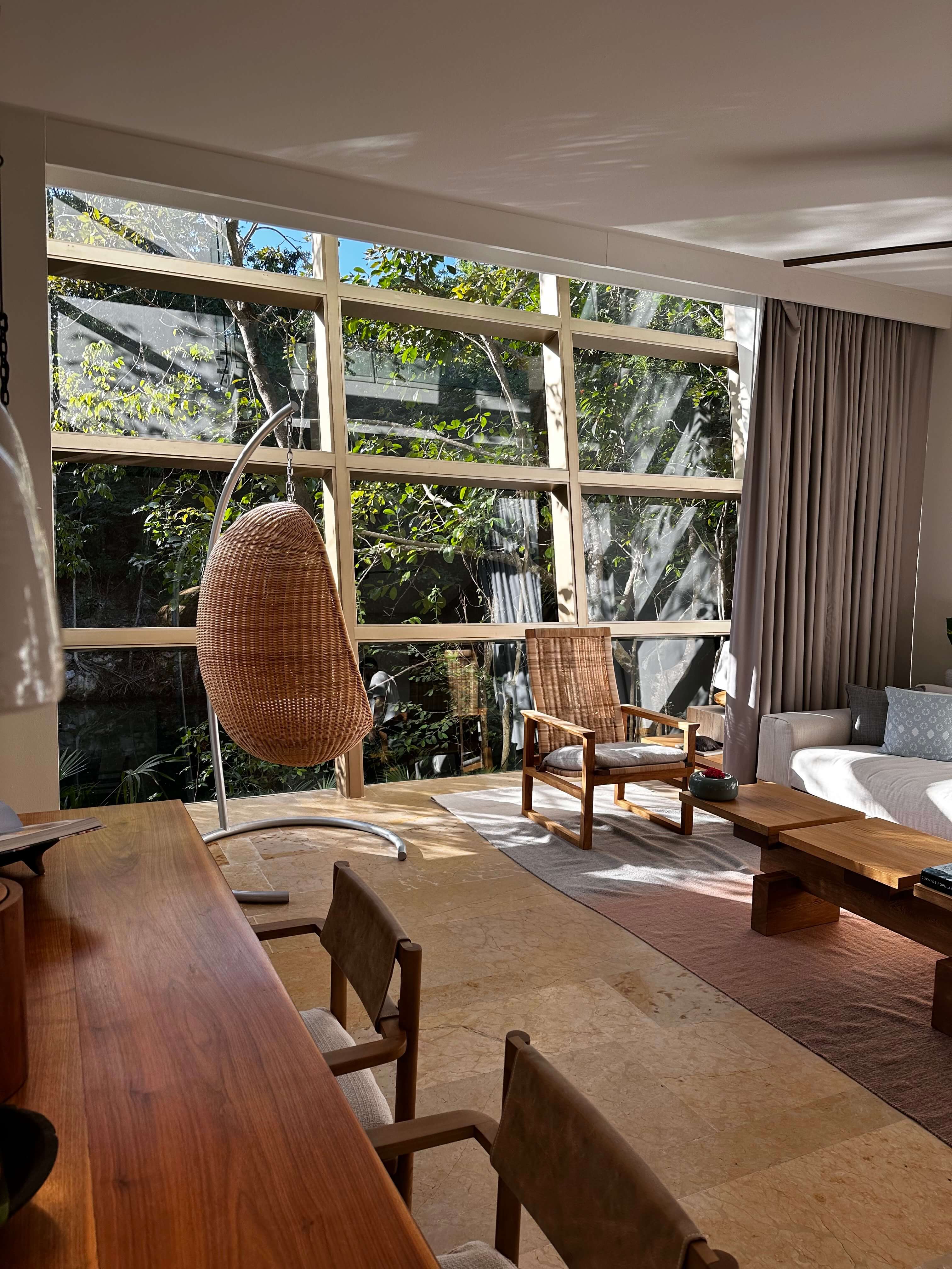 Rosewood Mayakoba, Mexico — Sunlit suite with woven hanging chair, wooden furnishings, and expansive glass doors opening to lush tropical gardens and cenote.