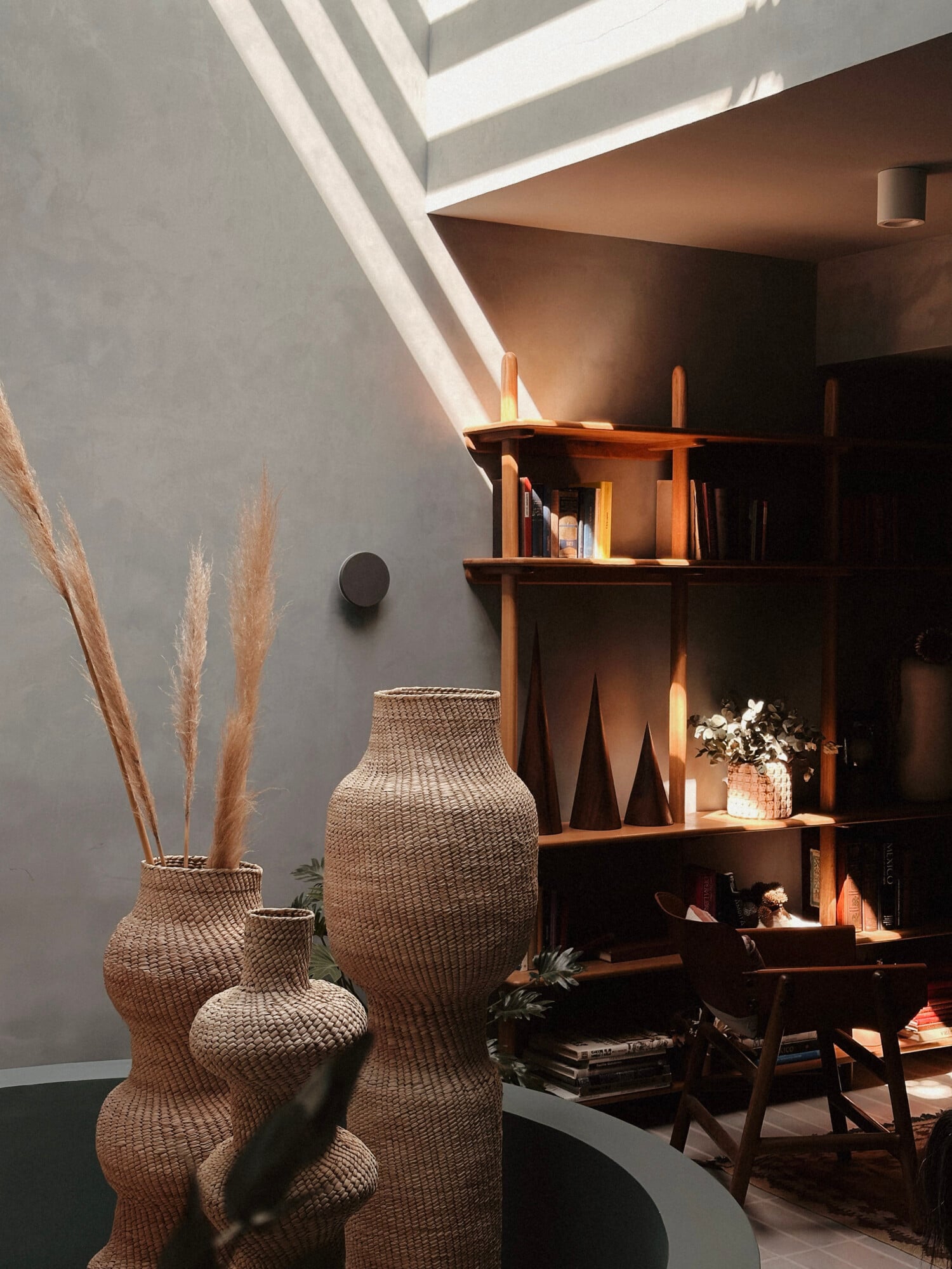 UMA Casa, Mexico — Minimalist interior with woven vessels, wooden shelving, pampas grass, and natural light from skylight casting geometric shadows on gray walls.