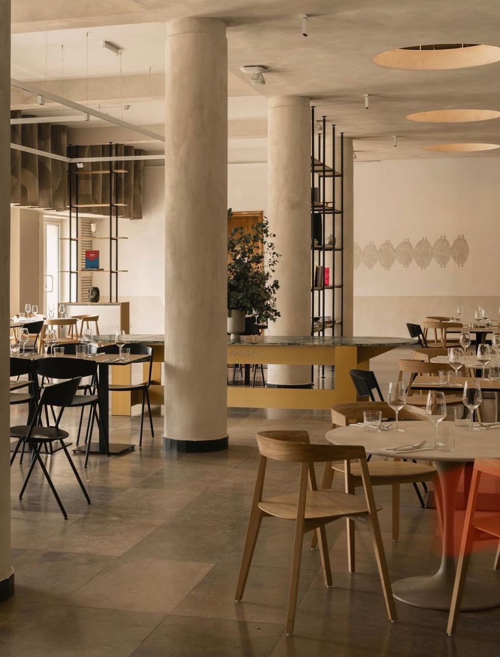 Muzealna, Europe — Minimalist restaurant interior with concrete columns, light wood furniture, polished stone flooring, and pendant lighting above dining tables.