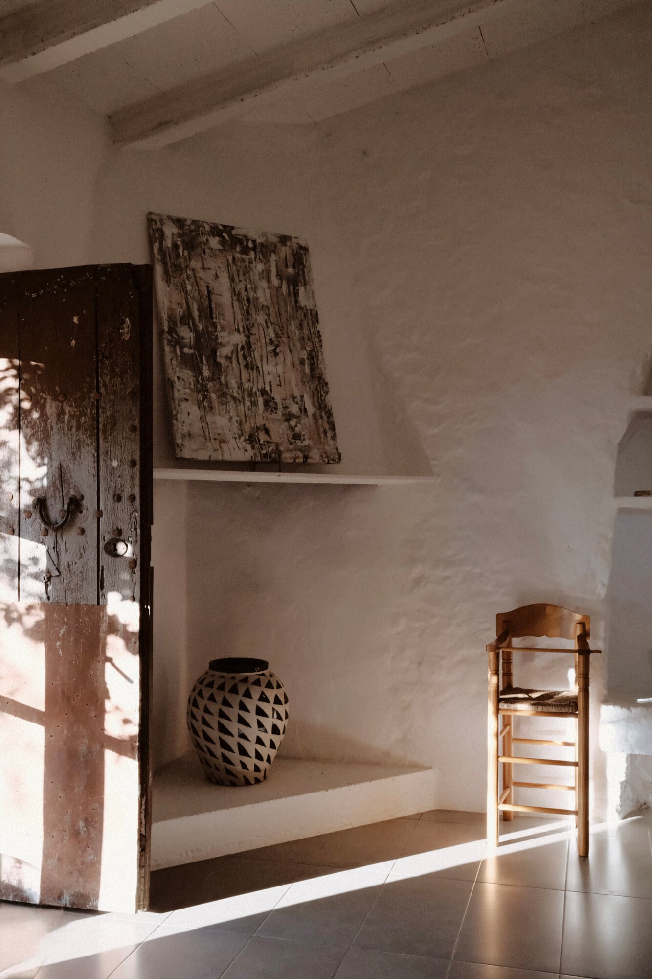 Vista Verde, Balearic Islands, Spain — Minimalist whitewashed interior with distressed wooden door, geometric patterned vessel, and woven chair casting shadows in natural light.