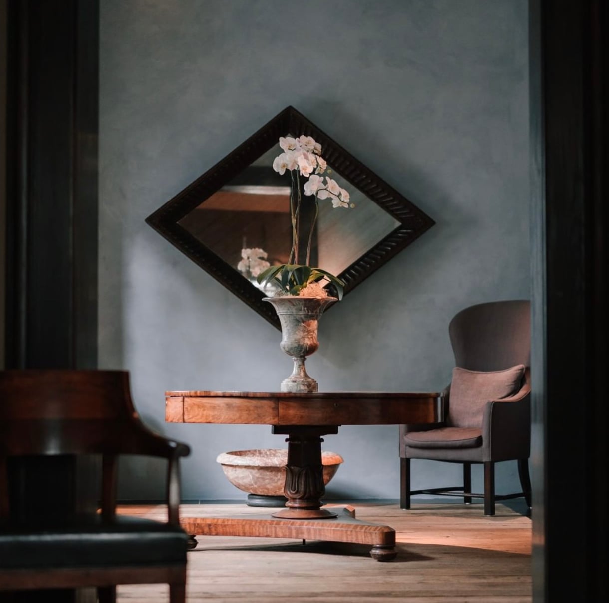 Purs, Europe — Minimalist dining room with orchid arrangement, diamond mirror, pedestal table, and upholstered chair bathed in natural light.