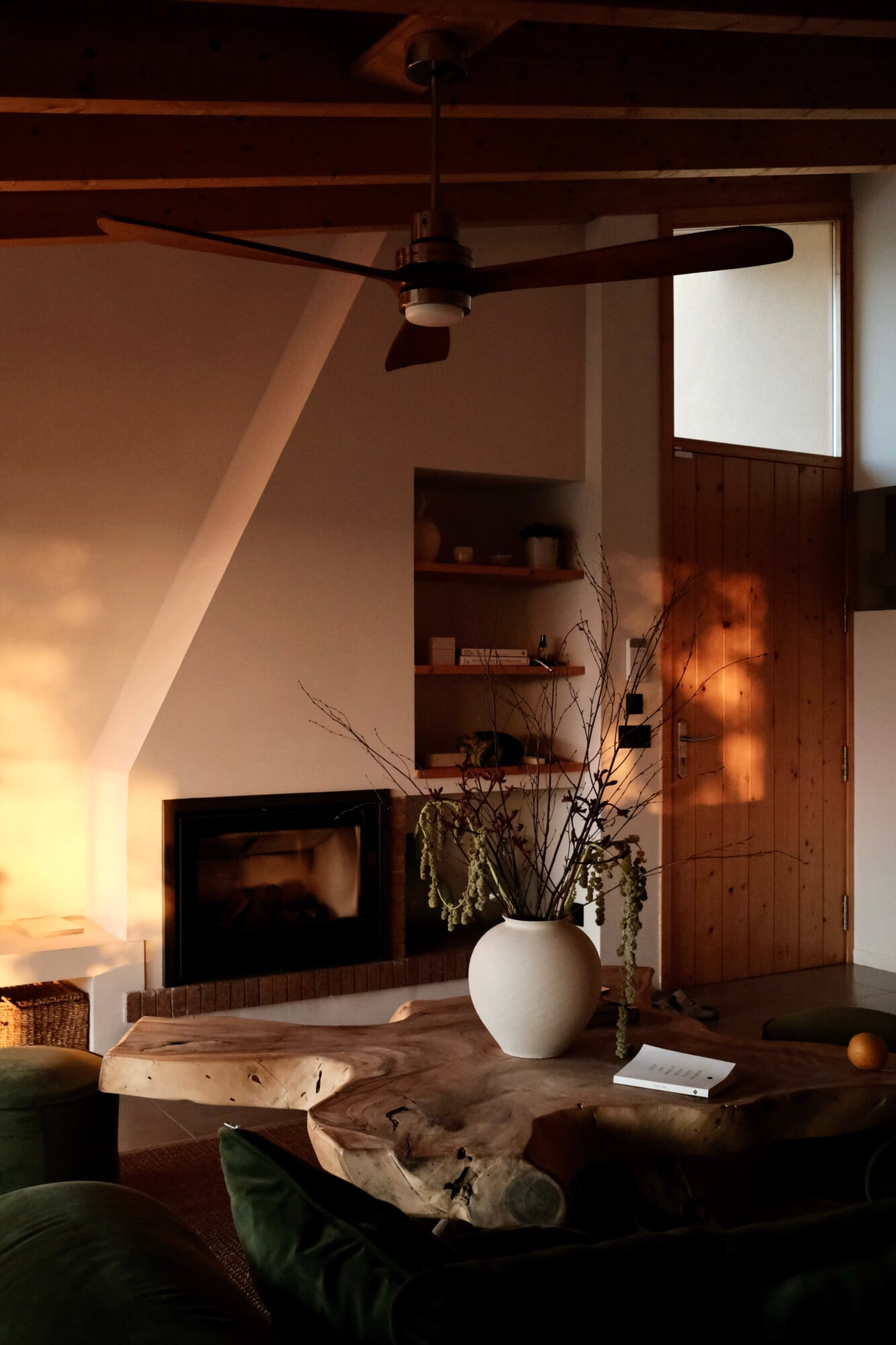 Vista Verde, Balearic Islands, Spain — Sunlit living room with exposed wooden beams, ceiling fan, fireplace, white ceramic vase with dried botanicals on driftwood table, built-in shelving, minimalist coastal design.