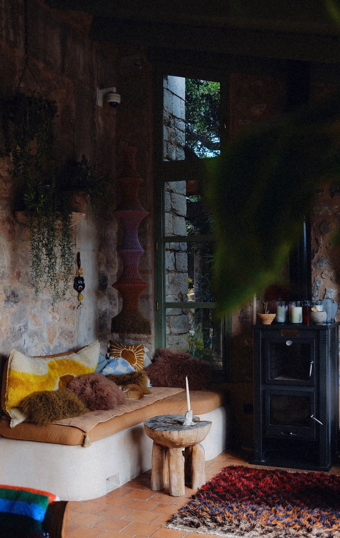 Hotel Corazón, Balearic Islands — Rustic stone interior with curved seating, woven textiles, wooden side table, terracotta floors, and natural light from tall windows.